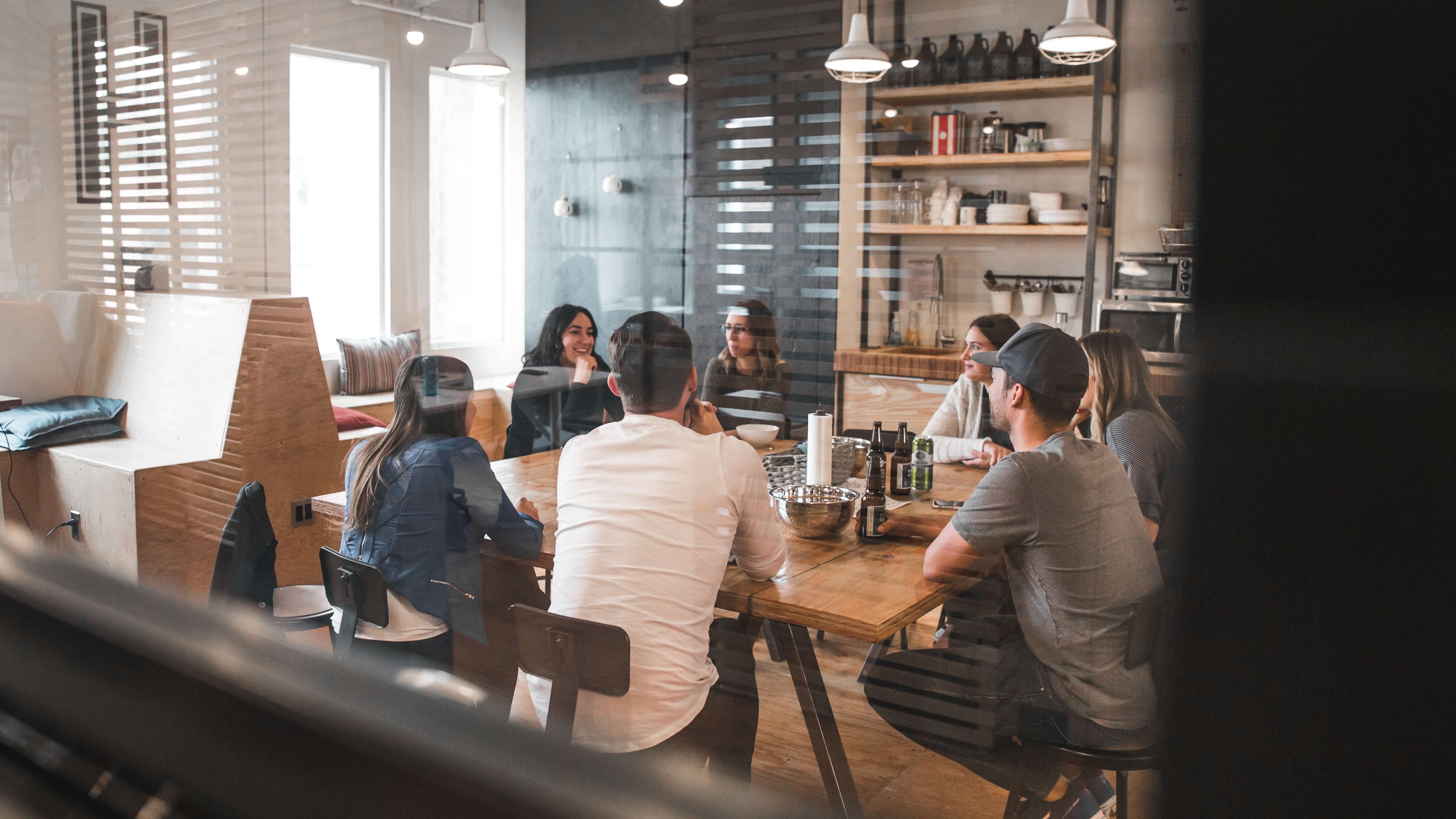 A imagem retrata um grupo de pessoas sentadas ao redor de uma mesa em um ambiente descontraído, que parece ser um espaço para refeições ou reuniões. O cenário sugere um espaço moderno, possivelmente urbano, com um design de interiores que inclui prateleiras expostas e uma variedade de garrafas e itens decorativos.
