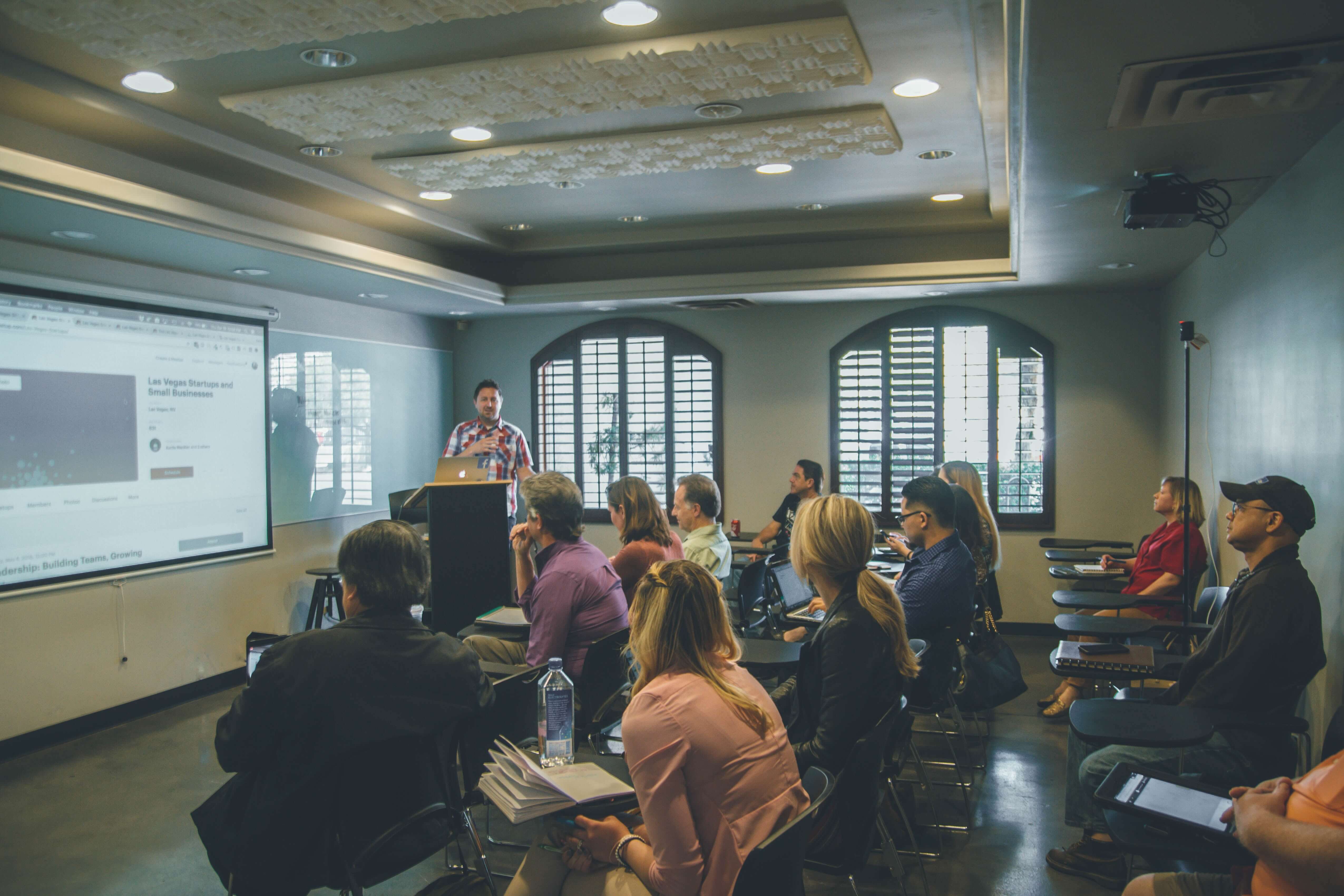 Sala de aula ou conferência com várias pessoas sentadas, atentas a uma apresentação. O apresentador está de pé à frente da sala, ao lado de um projetor que exibe informações em uma tela grande. A cena sugere um ambiente de aprendizado ou compartilhamento de informações, com os participantes engajados e interativos.