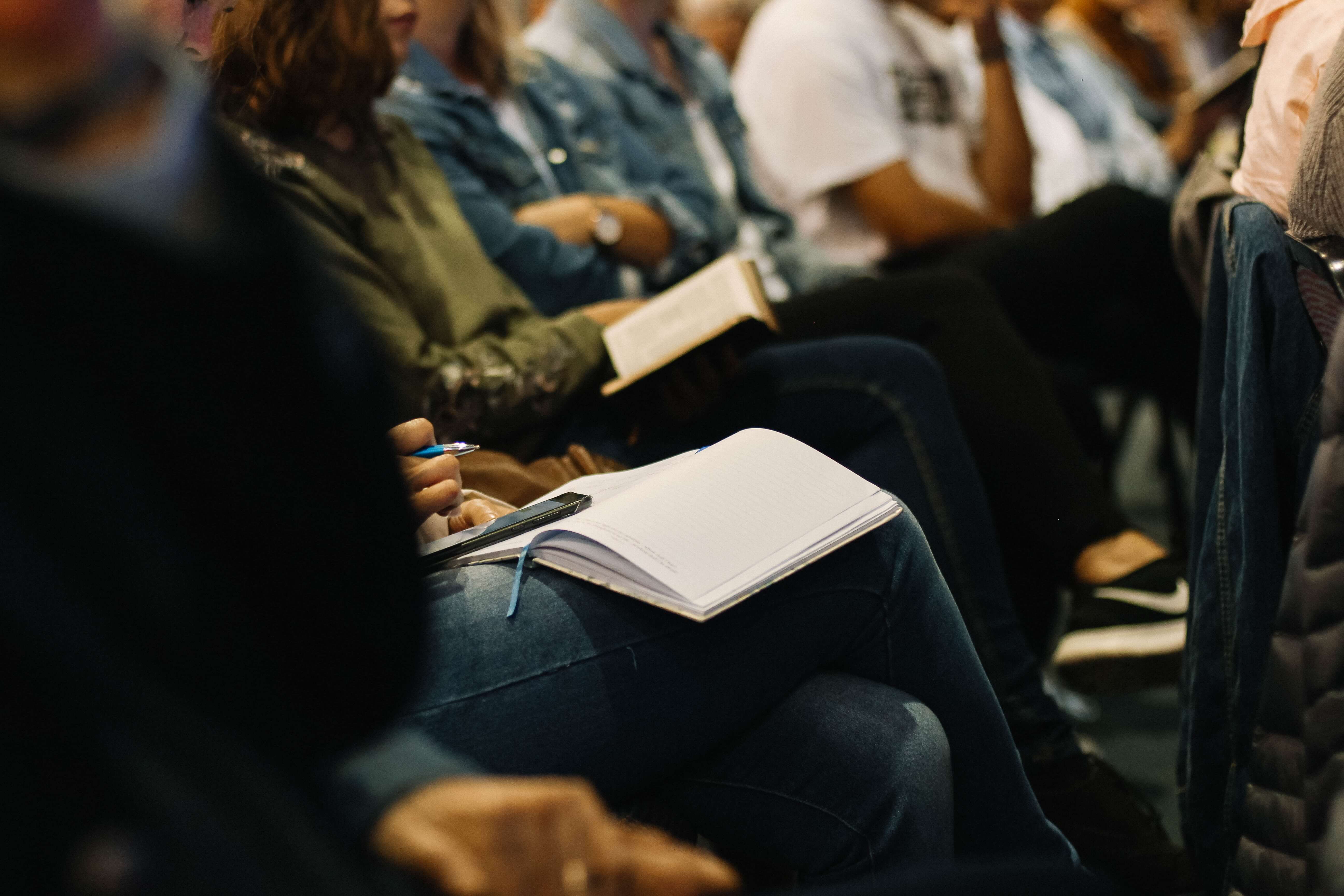 Ambiente de aprendizado ou conferência, com várias pessoas sentadas em fileiras. O foco está em uma pessoa segurando um caderno aberto e uma caneta, indicando que estão envolvidas em atividades de anotações e aprendizado.
