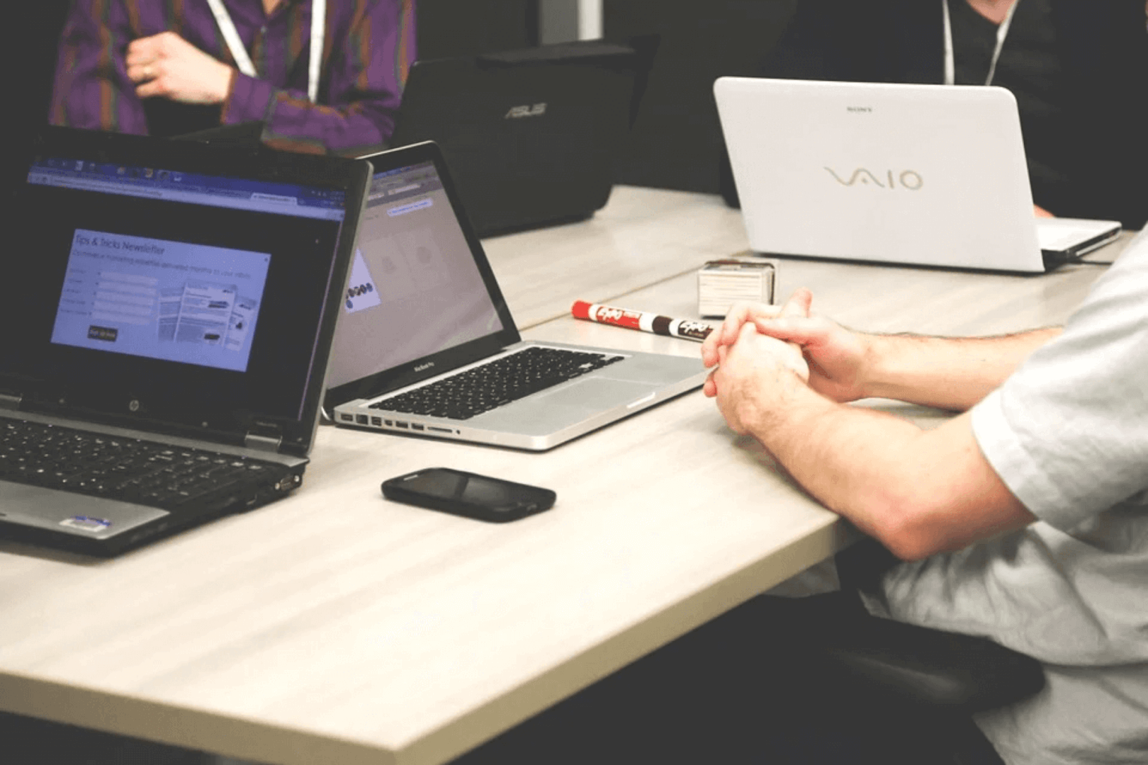 Reunião ou encontro onde as pessoas estão usando laptops. Um indivíduo tem as mãos cruzadas sobre a mesa, e há um smartphone e um marcador ao lado. A cena destaca a integração da tecnologia em ambientes colaborativos.