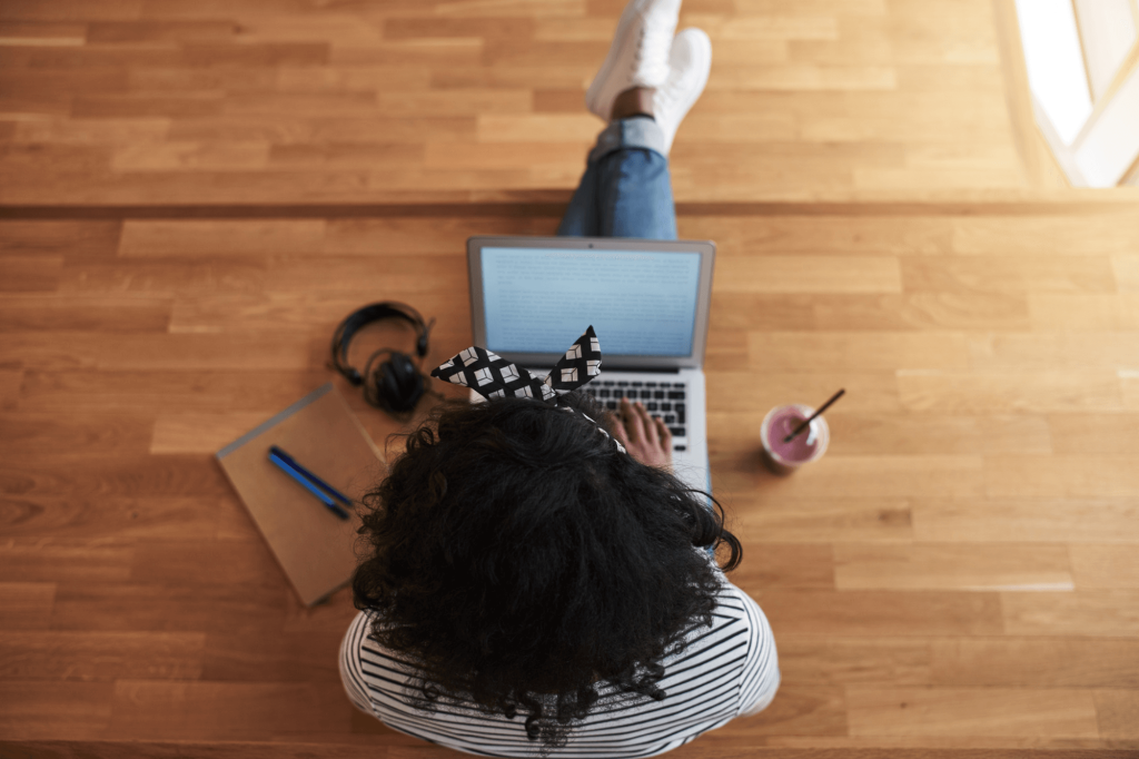 Pessoa sentada no chão, usando um notebook em um ambiente com piso de madeira clara. A perspectiva é de cima, mostrando a pessoa de costas enquanto digita no computador. Ela usa uma faixa ou laço preto e branco no cabelo, e veste uma camiseta listrada. Suas pernas estão esticadas à frente, e ela usa tênis brancos.