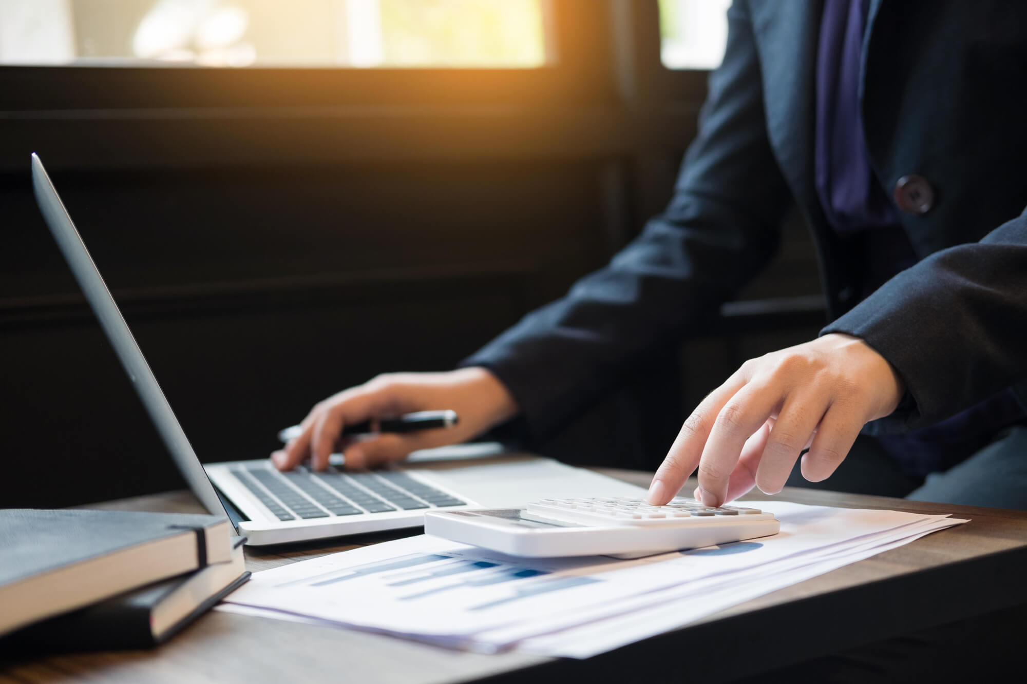 Pessoa em traje formal, possivelmente em um ambiente de escritório. A pessoa está usando um laptop que está aberto e ligado. Uma mão está no teclado do laptop e a outra mão está usando uma calculadora. Há documentos com gráficos visíveis espalhados na mesa perto do laptop. O ambiente é iluminado por luz natural vinda de uma janela ao fundo