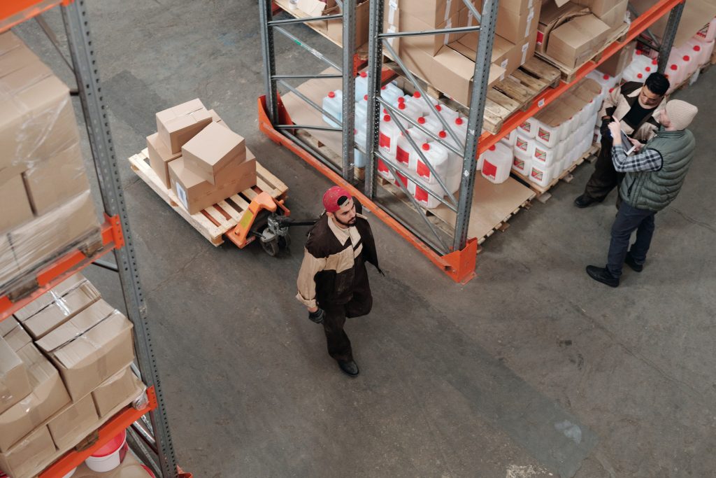 Ambiente de armazém interno com prateleiras altas repletas de várias caixas e mercadorias. Três funcionários estão fazendo o balanço de estoque. A perspectiva é de cima.