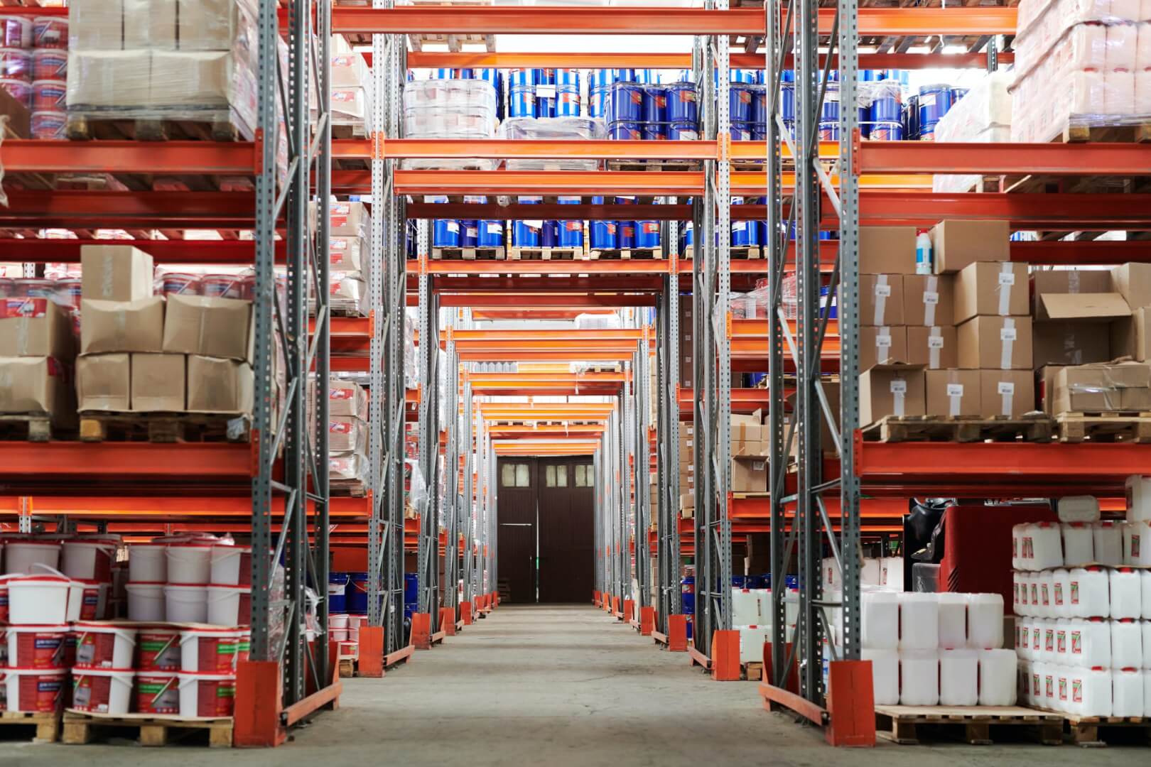 Interior de um grande depósito ou armazém. As prateleiras de metal altas estão organizadas em fileiras paralelas, criando corredores. Diversos produtos estão estocados nas prateleiras.