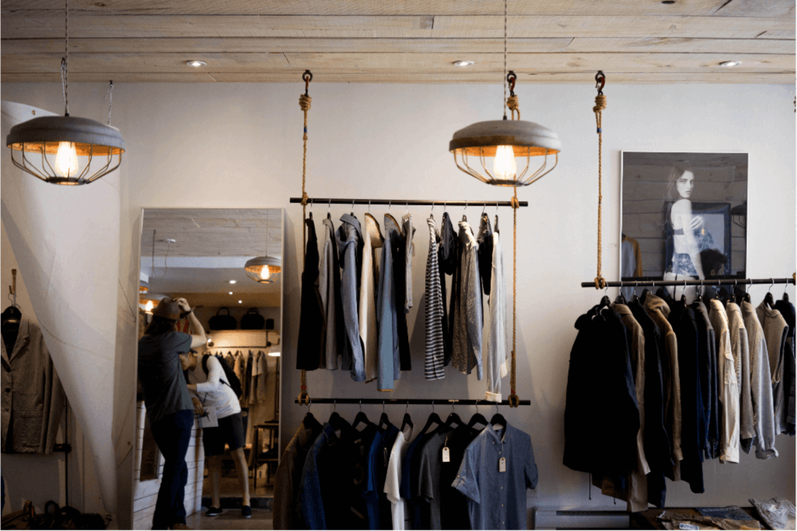 Interior de uma loja de roupas. Há dois lustres pendentes com design vintage, proporcionando uma iluminação suave ao espaço. Na frente, um rack de roupas exibe camisas e jaquetas em cores neutras.