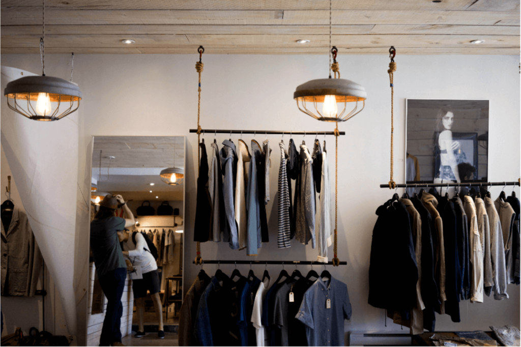 Interior de uma loja de roupas. Há dois lustres pendentes com design vintage, proporcionando uma iluminação suave ao espaço. Na frente, um rack de roupas exibe camisas e jaquetas em cores neutras. Oferta e demanda.