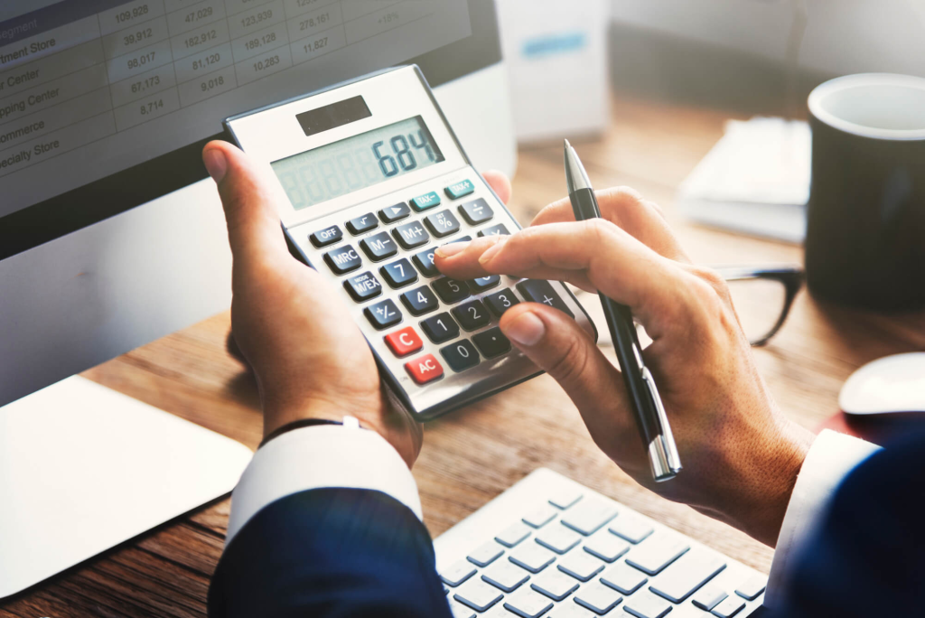 Mãos de uma pessoa usando calculadora sobre a mesa. A mão direita segura uma caneta, enquanto a esquerda pressiona teclas. Ao fundo, um monitor exibe planilha com dados e gráficos. Representa o cálculo de markup.