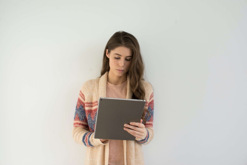 Pessoa segurando um tablet com ambas as mãos. A pessoa está vestindo uma blusa multicolorida e parece estar em pé contra um fundo claro e simples