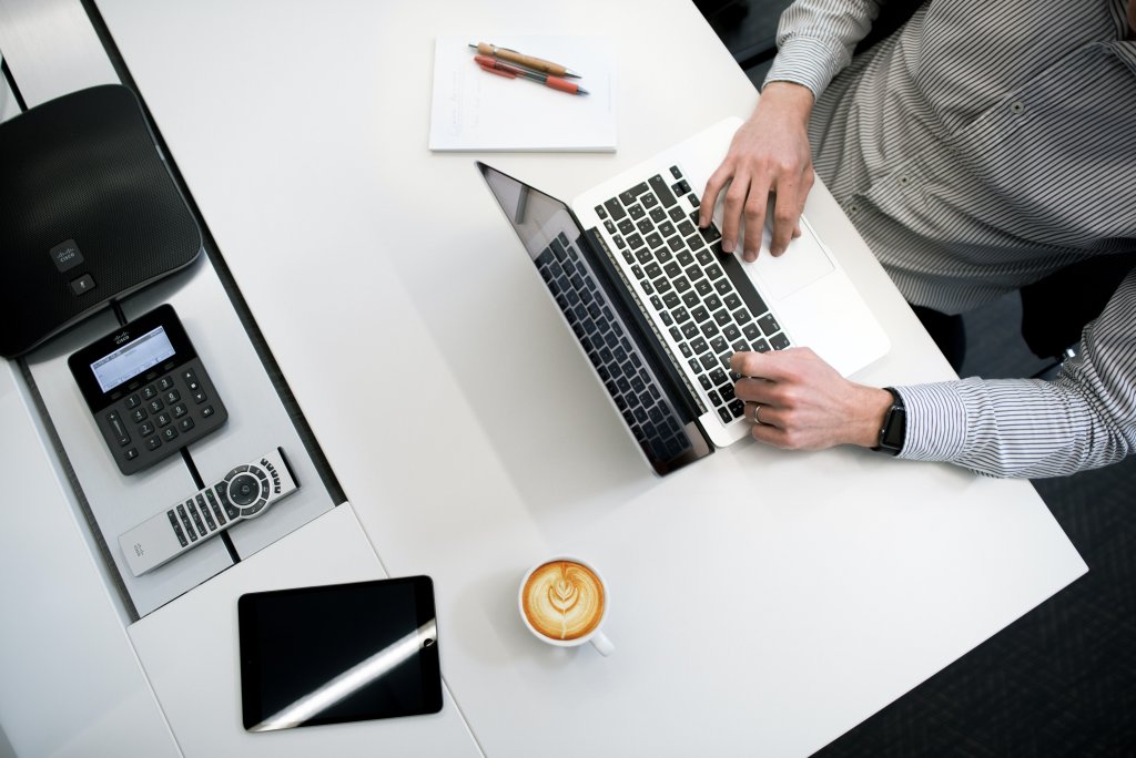 Pessoa trabalhando em um laptop sobre mesa branca. À esquerda, bloco de notas, canetas e calculadora preta. À direita, xícara de café com arte de latte. Smartphone está virado para baixo.