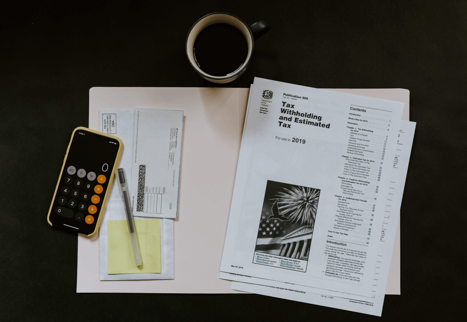 Vários itens sobre superfície escura: folha de papel branca, copo preto de café, smartphone com capa amarela e preta, duas canetas, adesivo amarelo, envelope, e folhas impressas. Representa o cálculo de markup.