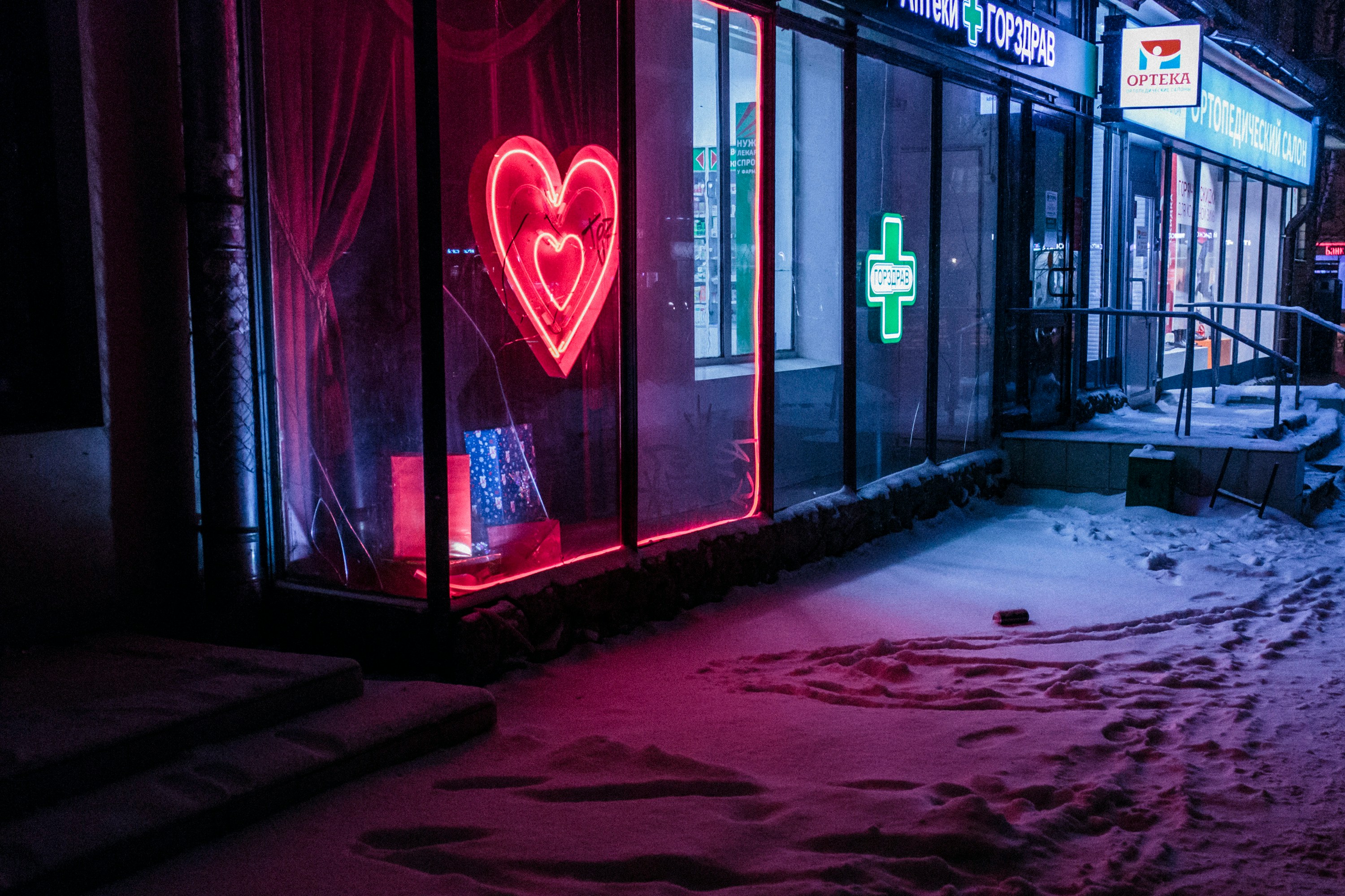 A imagem apresenta uma cena noturna de rua com neve no chão. Há uma loja com grandes vitrines de vidro, iluminadas por letreiros de neon. Um dos letreiros de neon tem a forma de um coração e brilha em vermelho, enquanto outro letreiro, que parece estar dentro da loja, brilha em verde e se assemelha a uma cruz médica. A iluminação desses letreiros projeta reflexos coloridos na calçada coberta de neve. A cena sugere que pode ser uma farmácia devido à presença do letreiro verde com a cruz, que é comumente associada a farmácias em muitos países.