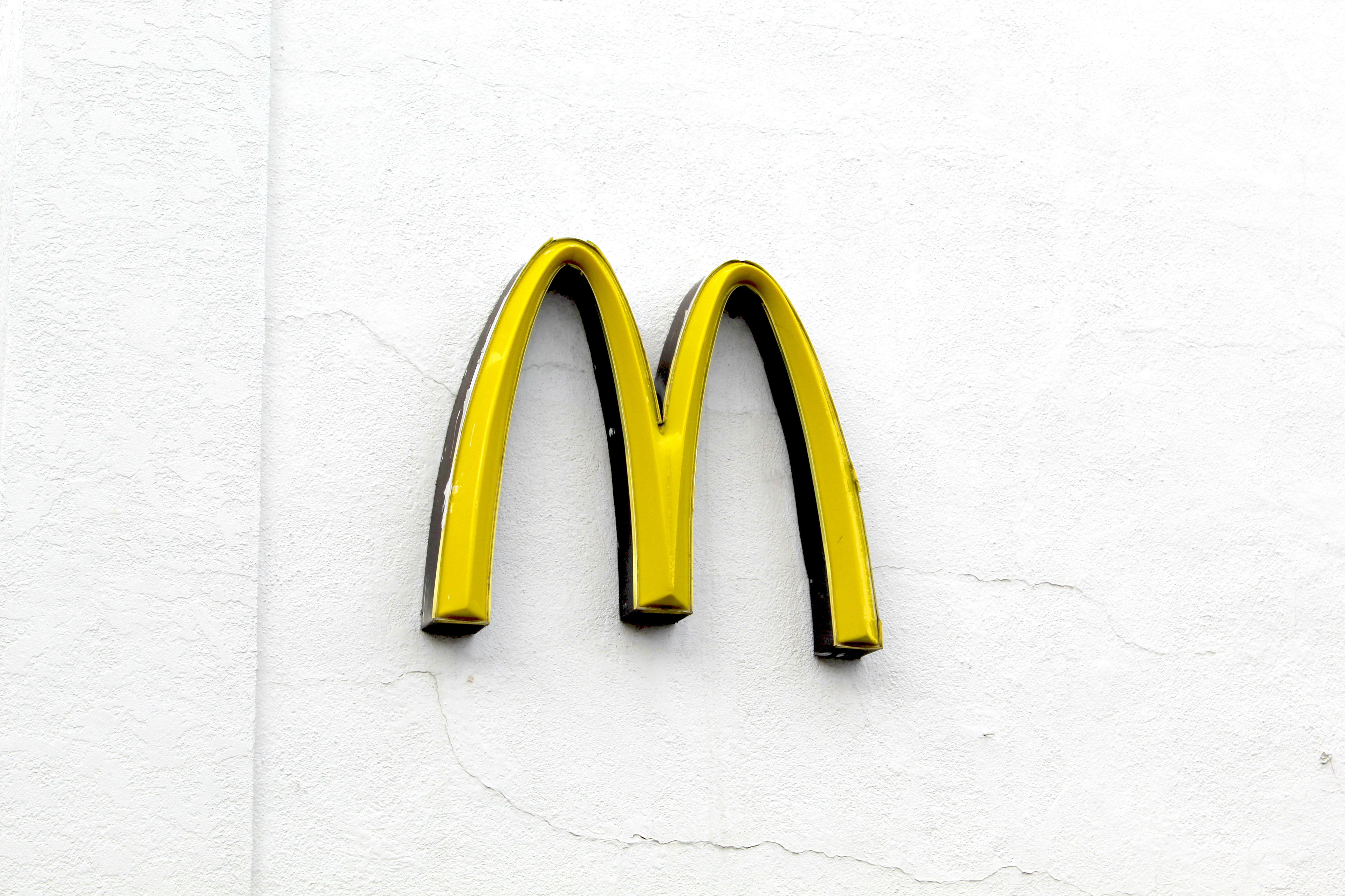 Icônico logotipo do McDonald’s, composto por dois arcos dourados entrelaçados que formam a letra “M”. O logotipo está em relevo, destacando-se contra um fundo branco liso. Sua cor vibrante e bordas pretas conferem uma identidade visual marcante. Não há outros elementos visíveis na imagem, concentrando todo o foco no símbolo mundialmente reconhecido.