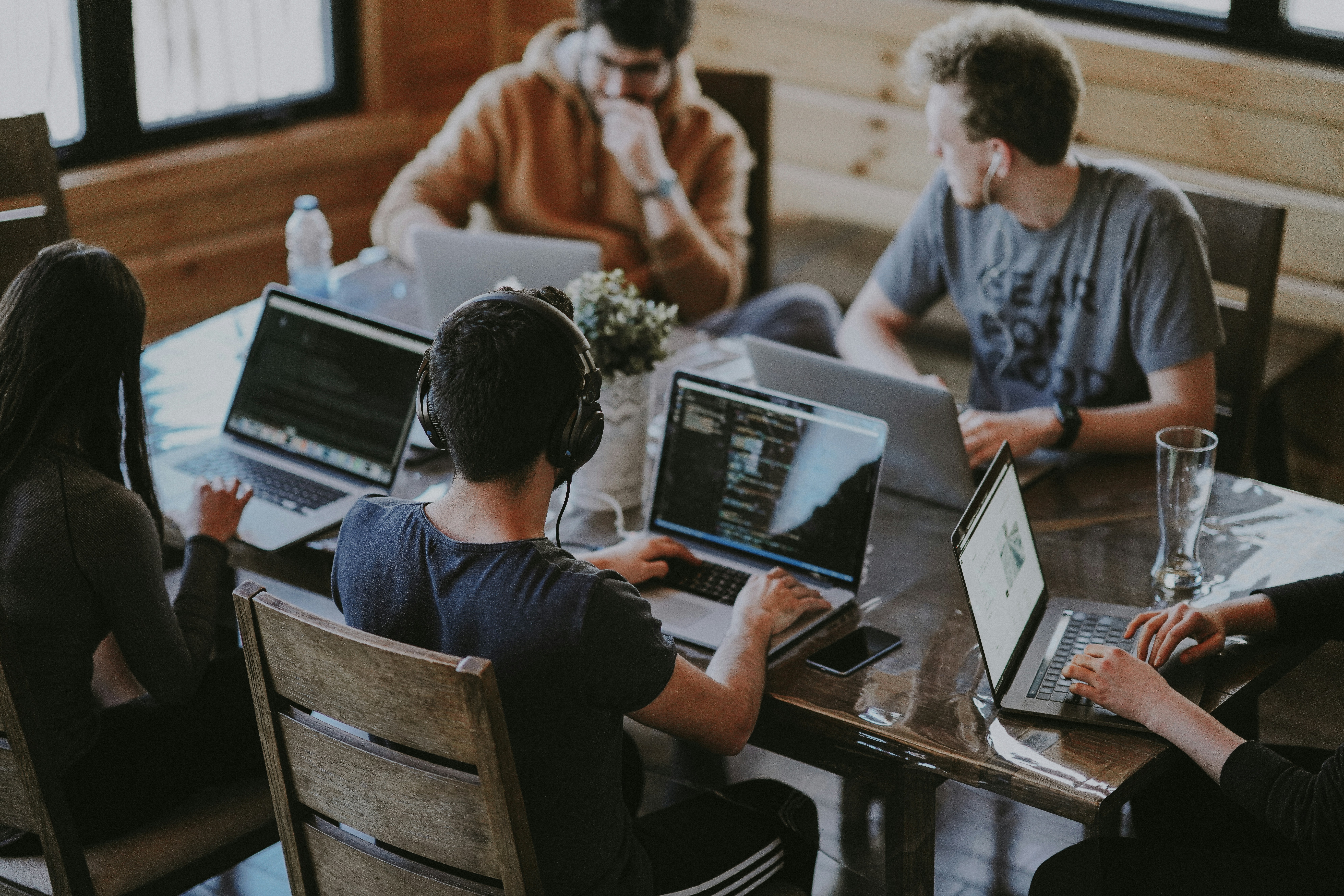 A imagem captura uma cena de quatro indivíduos sentados ao redor de uma mesa de madeira, cada um concentrado em seu laptop. O ambiente parece ser uma sala aconchegante e bem iluminada, com interiores de madeira.