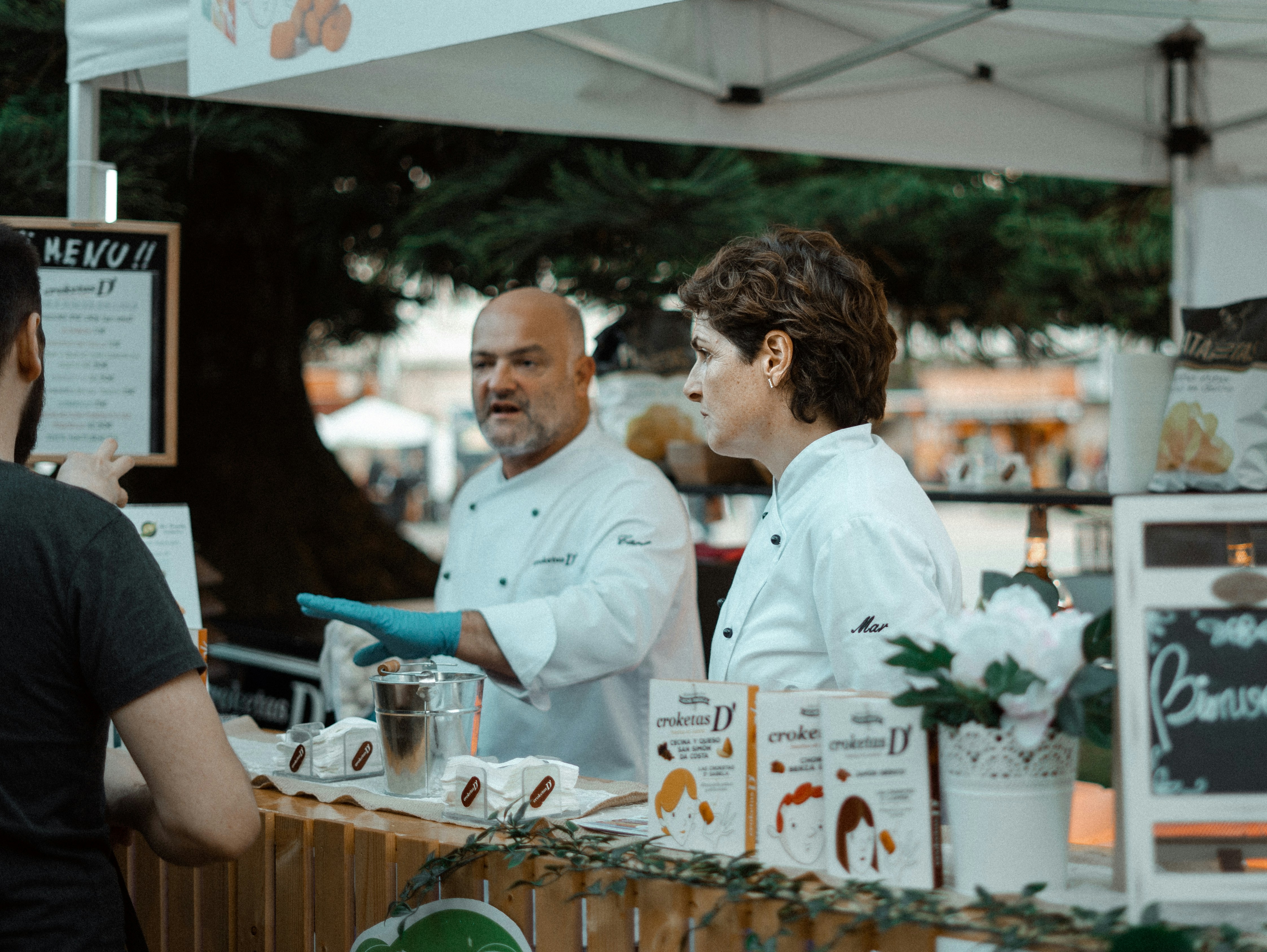 Dois chefs em jalecos brancos atendendo um cliente em uma barraca de comida ao ar livre. Os chefs estão vestindo luvas azuis e exibem produtos embalados na frente da barraca. O menu está pendurado no canto superior esquerdo, mas o texto não é claramente visível. A cena ocorre em um ambiente ao ar livre, possivelmente durante um evento ou feira, com outras barracas e pessoas ao fundo.