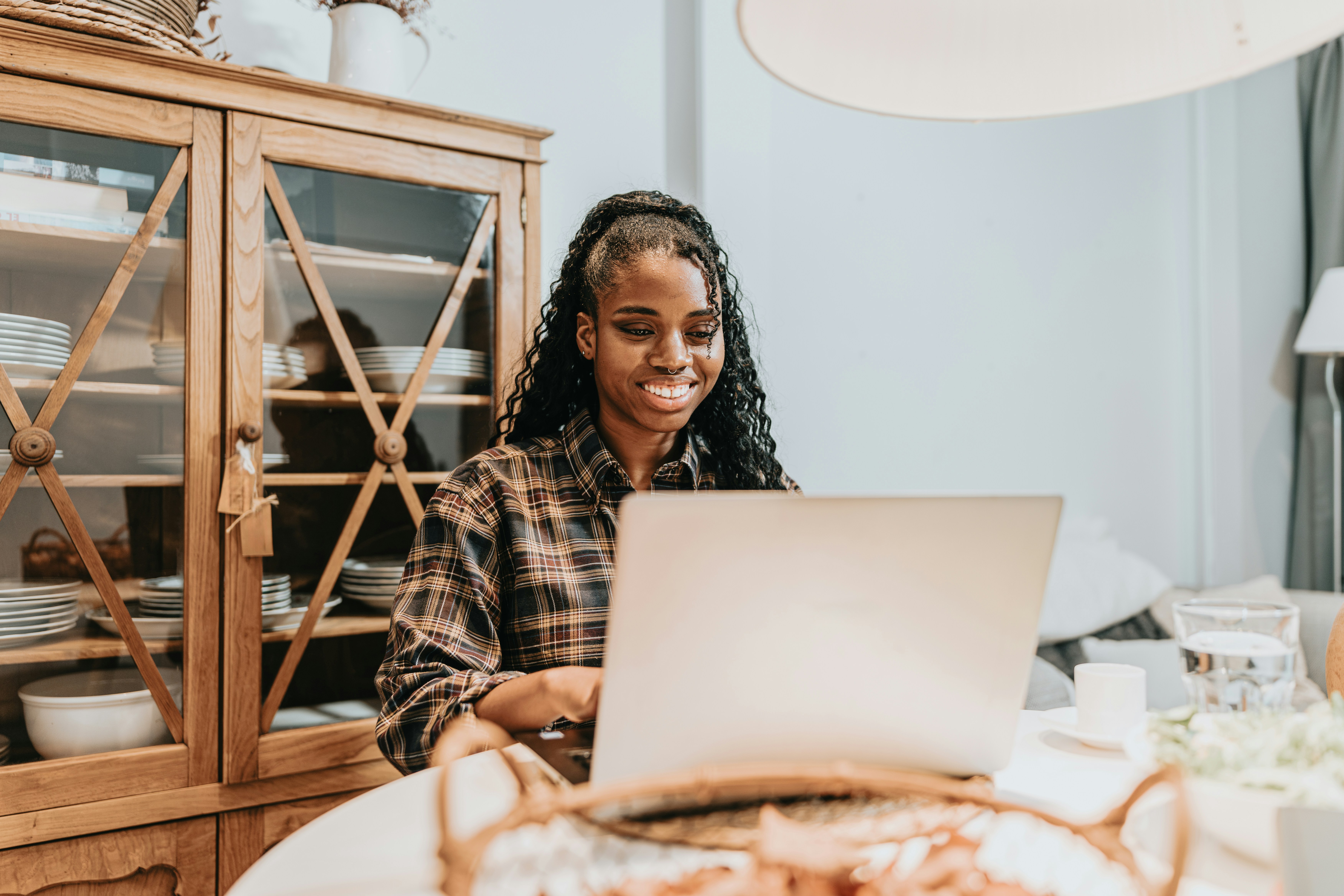 Pessoa sentada em frente a um laptop aberto sobre uma mesa abrindo um MEI para e-commerce. A pessoa parece estar envolvida em trabalho ou estudo, indicado pela presença do laptop. O cenário parece ser um ambiente doméstico, com móveis como um armário de madeira com portas de vidro e diversos itens decorativos dentro dele. Uma luminária com uma grande cúpula está visível na mesa, fornecendo iluminação. A cena captura um cenário comum nos dias atuais, de alguém trabalhando em casa ou remotamente, o que é relevante dada a tendência atual de teletrabalho e educação online.