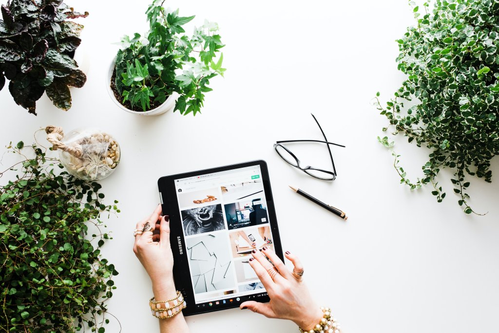 Vista de cima das mãos de uma pessoa segurando um tablet, que parece estar aberto na página do Pinterest, exibindo várias imagens. Ao redor do tablet, há várias plantas em vasos com folhagem verde exuberante, um par de óculos e uma caneta sobre uma superfície branca.