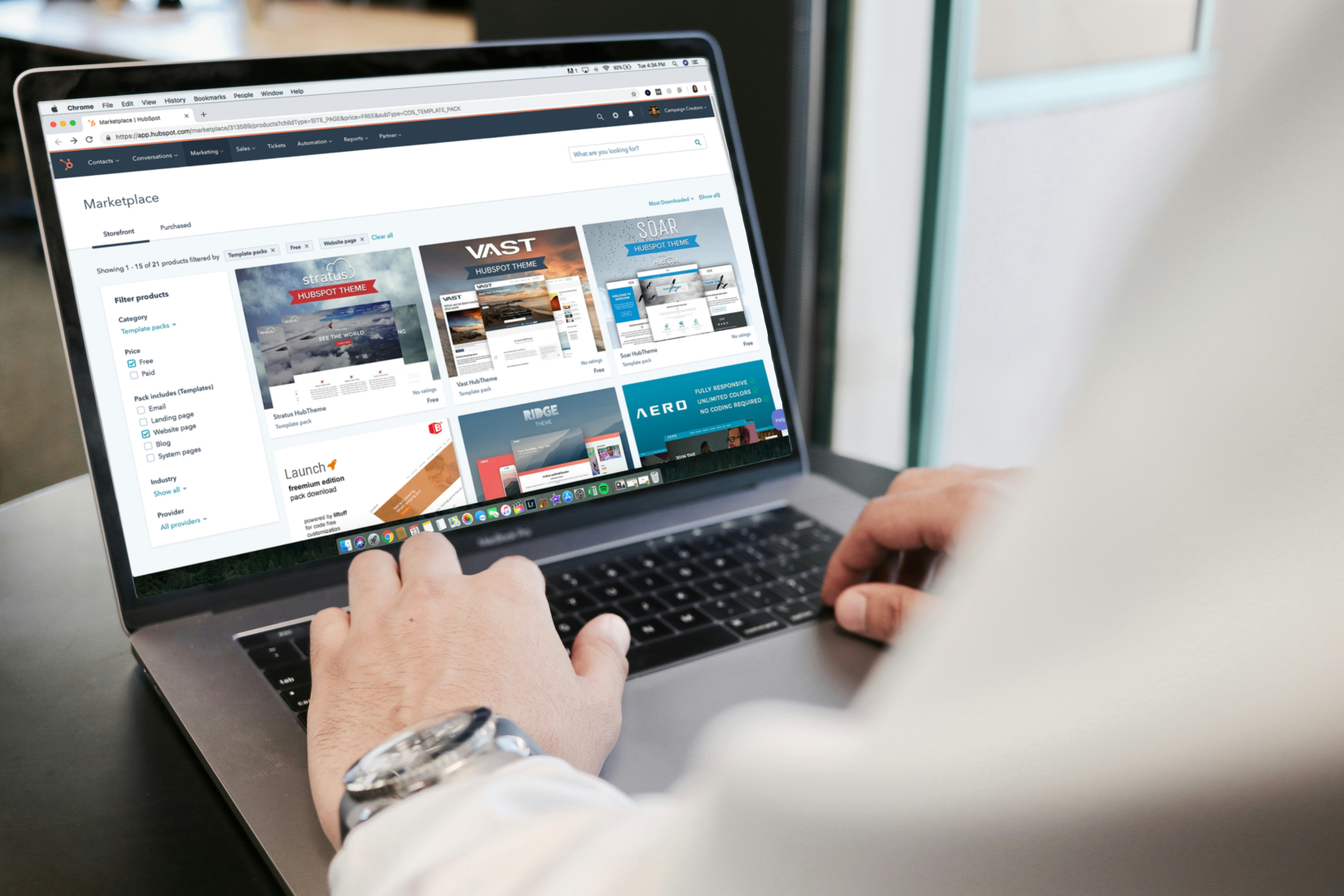 A imagem mostra uma visão de ombro de uma pessoa usando um laptop. A tela do laptop exibe o que parece ser um site com o título “Marketplace” no topo. Diferentes seções, como “Top picks”, “Categorias” e “Popular”, são visíveis na página. A interface do usuário inclui imagens e texto descrevendo diferentes serviços ou produtos, como imóveis e modelos de design. A pessoa usando o laptop veste uma camisa branca e um relógio no pulso esquerdo, sugerindo que ela pode estar em um ambiente profissional ou de trabalho. Essa imagem destaca a importância de abrir um e-commerce.