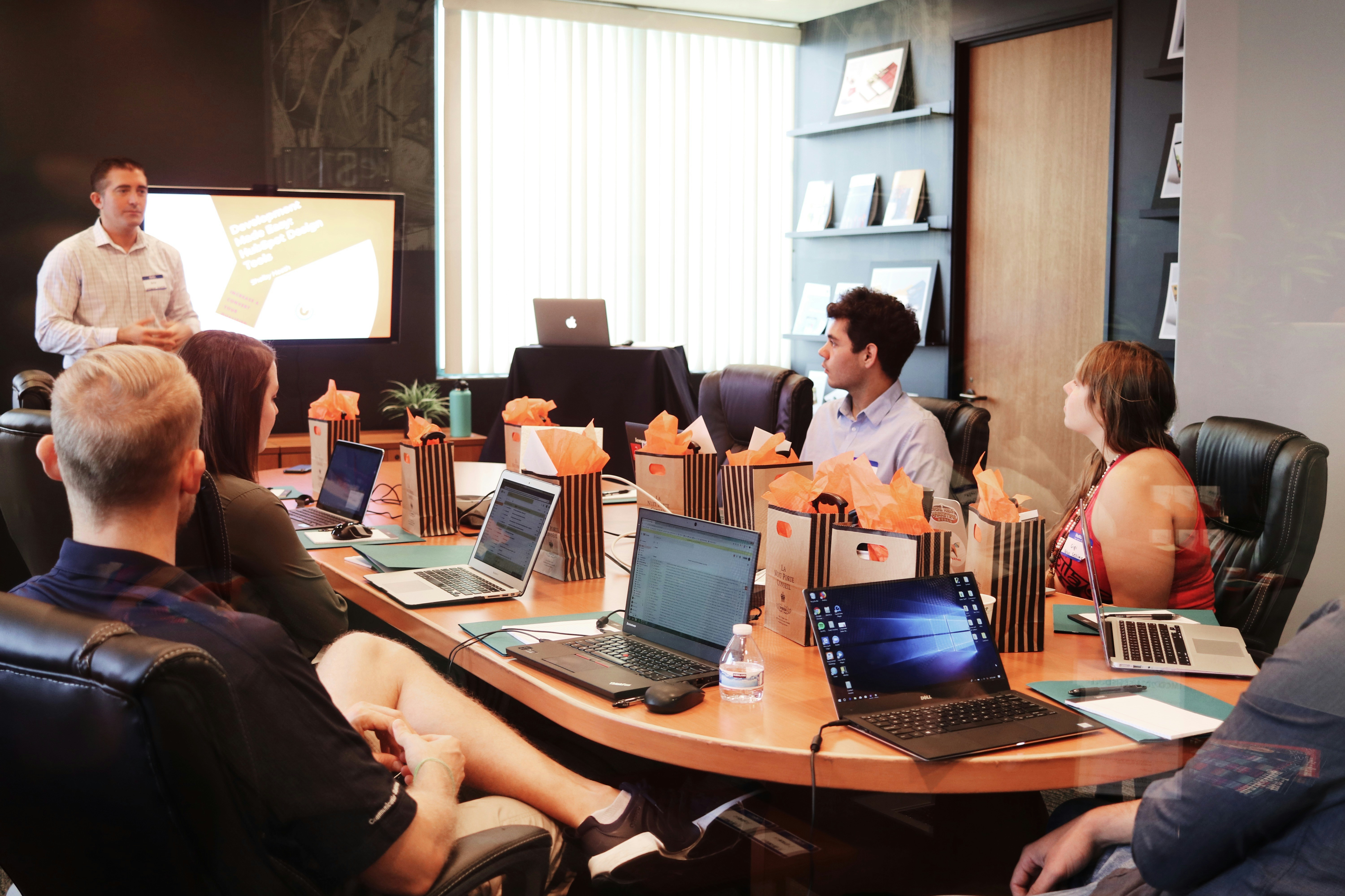 Reunião em um escritório moderno, com pessoas sentadas ao redor de uma mesa oval, focadas em um apresentador próximo a uma tela de projeção. A sala é bem iluminada e decorada com prateleiras de livros ao fundo. Vários laptops estão abertos sobre a mesa, e sacolas de papel marrom estão à frente de cada pessoa