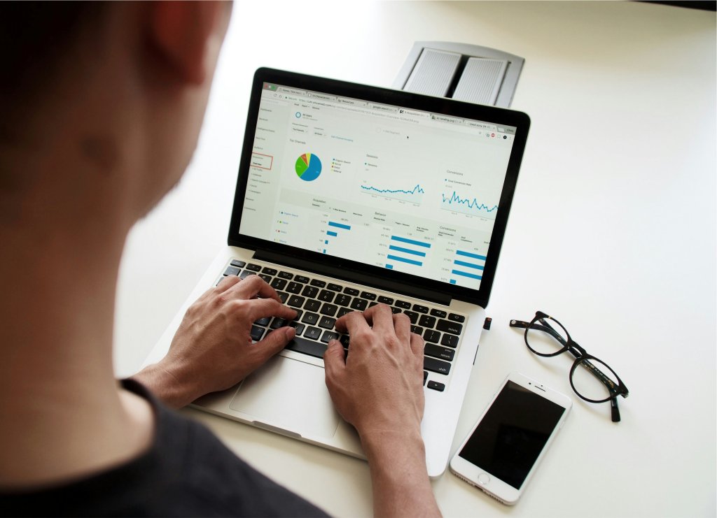 A imagem mostra uma visão de ombro de uma pessoa usando um laptop que exibe vários gráficos e gráficos indicando alguma forma de análise de dados ou métricas de negócios. O laptop está sobre uma superfície branca, e ao lado dele estão um par de óculos e um smartphone.