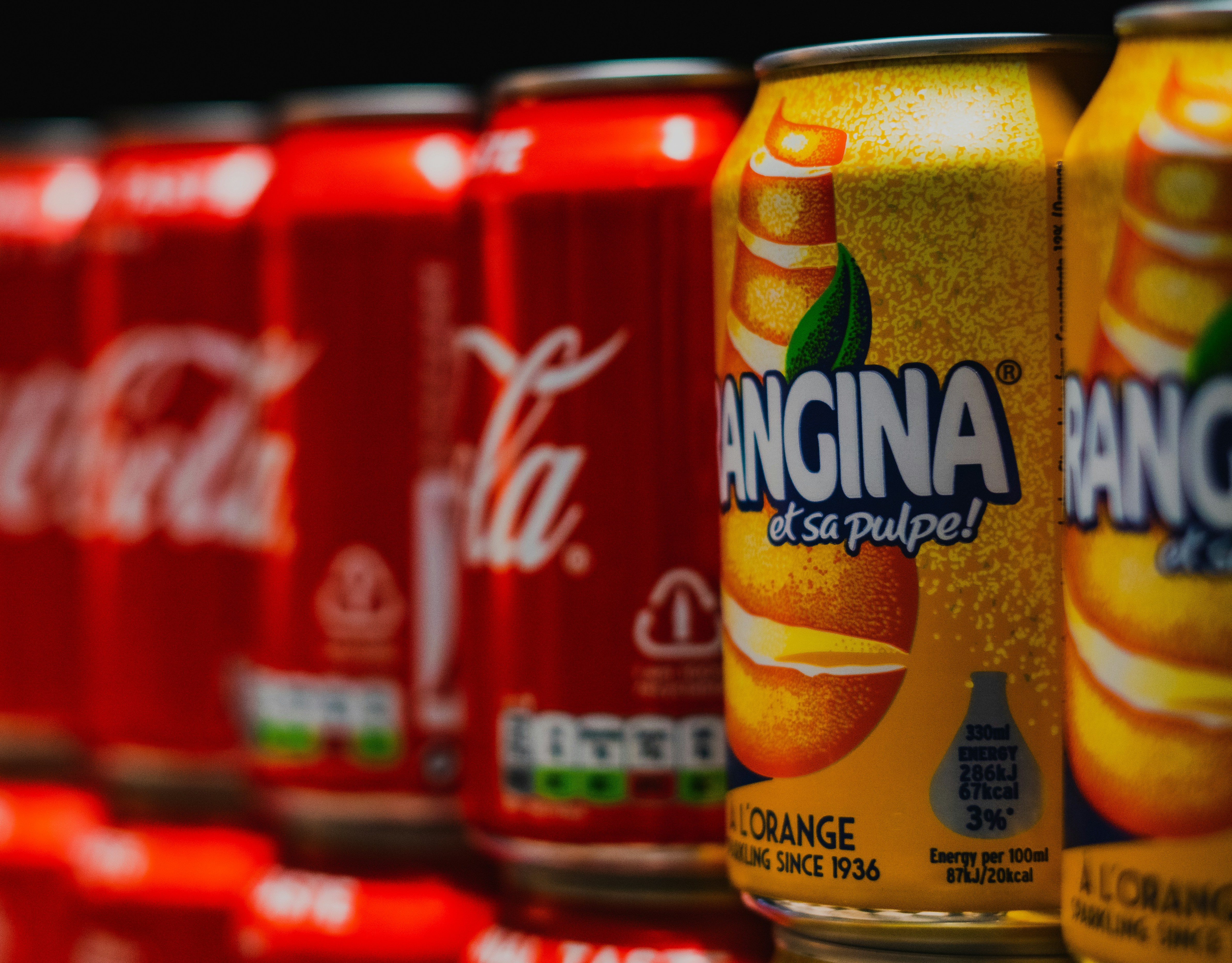  A imagem exibe uma coleção de latas de bebidas em close-up, com foco em uma lata central amarela da marca “ORANGINA”. A lata destaca a frase “avec sa pulpe!” sugerindo que contém polpa. As latas ao redor são vermelhas, provavelmente de cola, com o icônico script branco da Coca-Cola. Essa combinação de Orangina vibrante com as clássicas latas de Coca-Cola cria um contraste interessante de cores e identidades de marca, possivelmente indicando uma escolha entre cola tradicional e sabores cítricos com polpa natural.