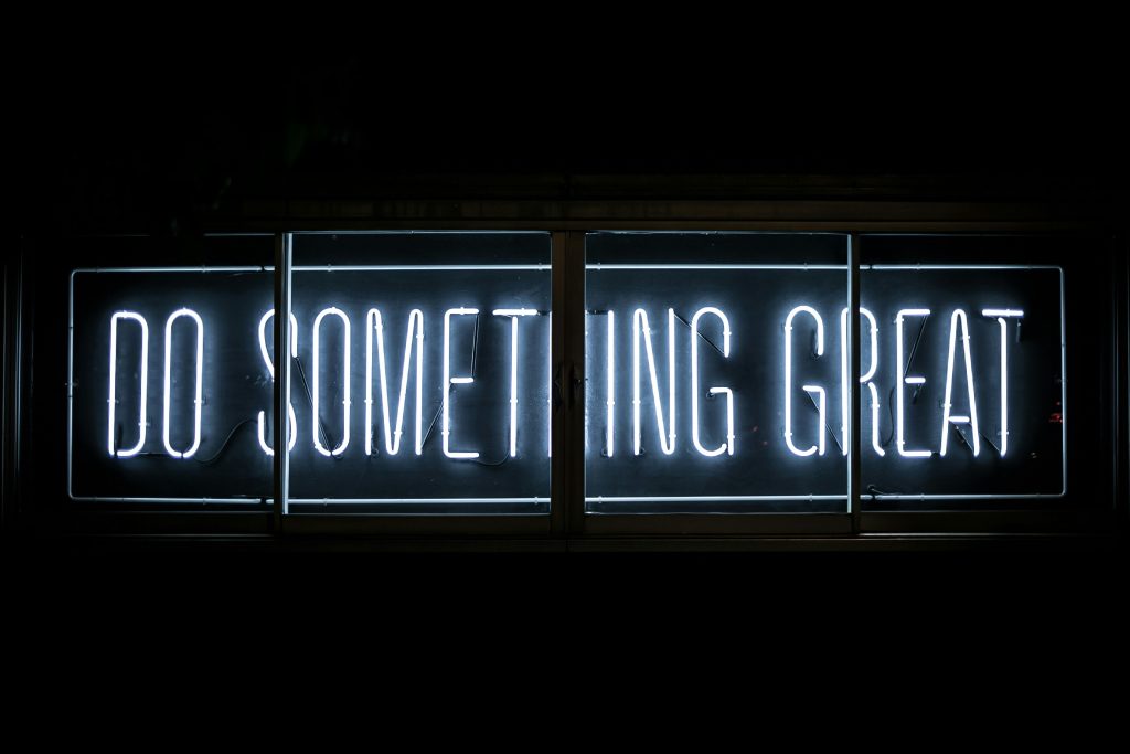 A imagem mostra uma placa de neon com a frase "DO SOMETHING GREAT" (em tradução livre, "FAÇA ALGO GRANDE"). A placa brilha em luz branca contra um fundo escuro, destacando-se de maneira impactante e inspiradora.