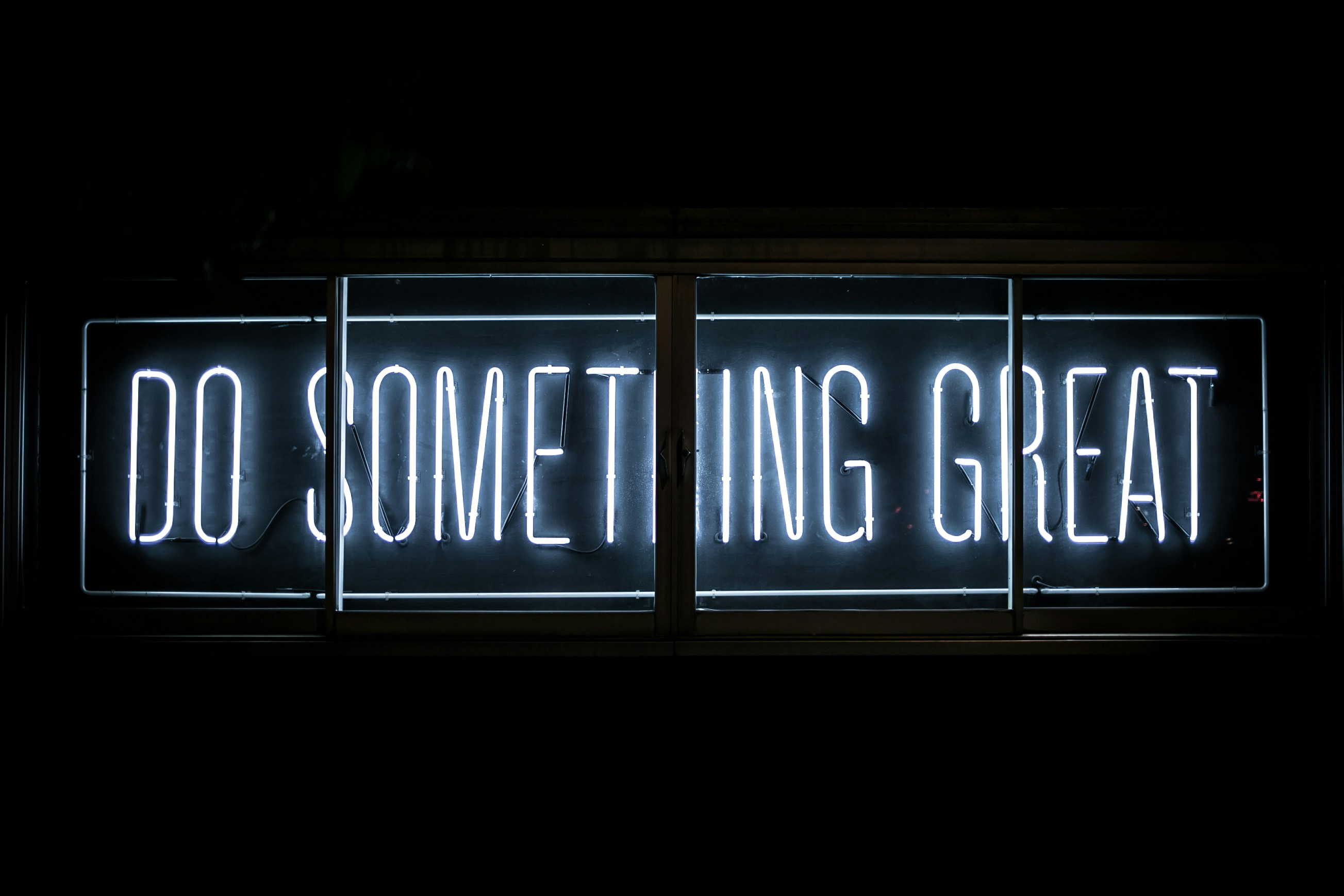 A imagem mostra uma placa de neon com a frase "DO SOMETHING GREAT" (em tradução livre, "FAÇA ALGO GRANDE"). A placa brilha em luz branca contra um fundo escuro, destacando-se de maneira impactante e inspiradora.
