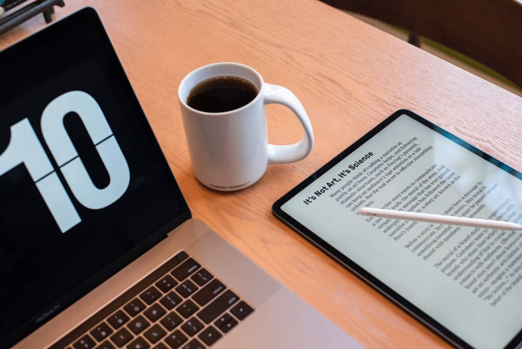 Ambiente de trabalho moderno com três itens principais: um laptop à esquerda com o número “10” exibido na tela, uma caneca de cerâmica branca cheia de café no centro, e um tablet à direita sendo usado com uma caneta stylus. A tela do tablet contém vários parágrafos de texto. A configuração está sobre uma superfície de madeira, possivelmente uma mesa, e há luz natural vindo do canto superior direito, sugerindo proximidade com uma janela.