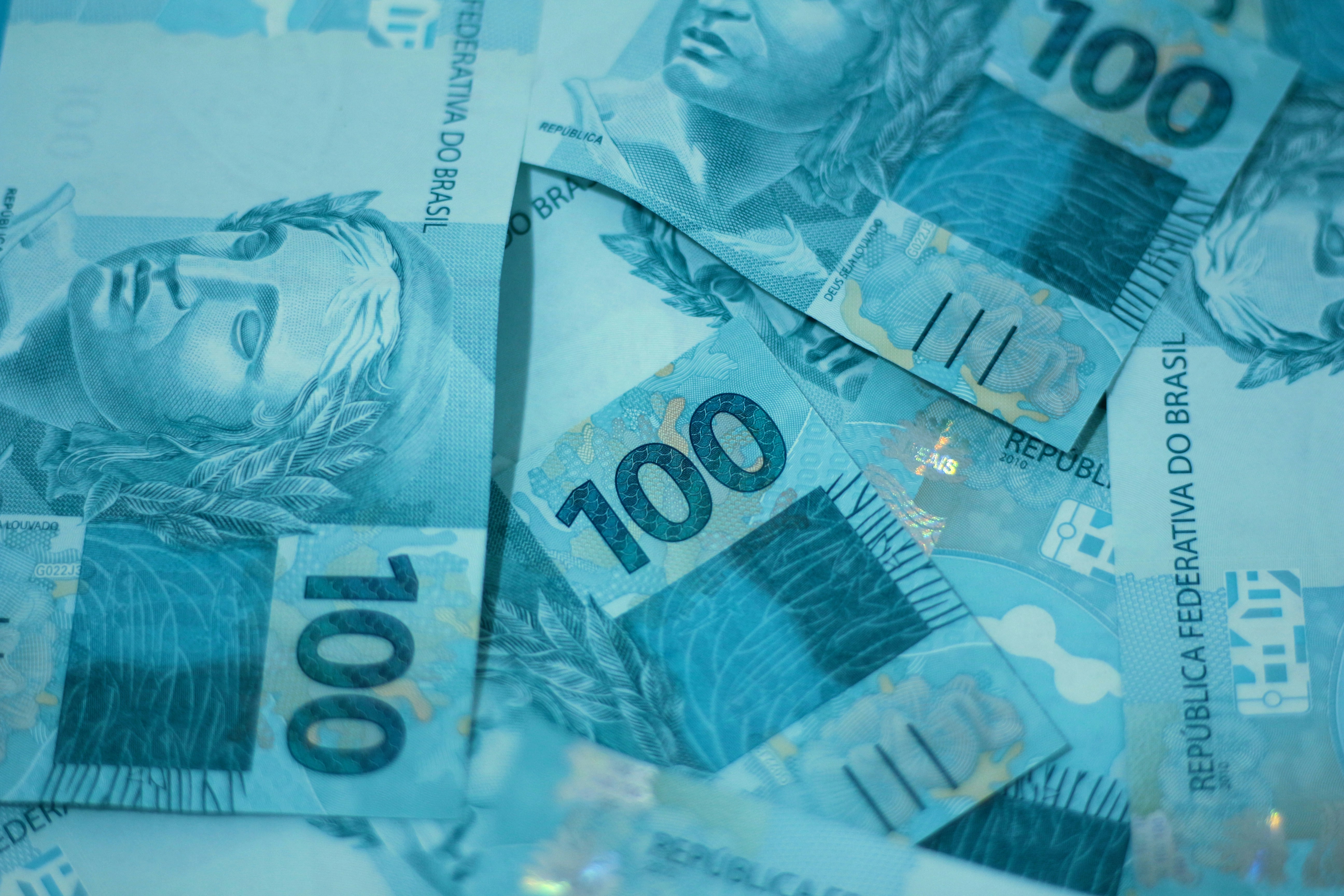A imagem mostra várias notas de 100 reais brasileiras. As cédulas estão sobrepostas e espalhadas, com destaque para a cor azul e a imagem da Efígie da República, um símbolo do Brasil