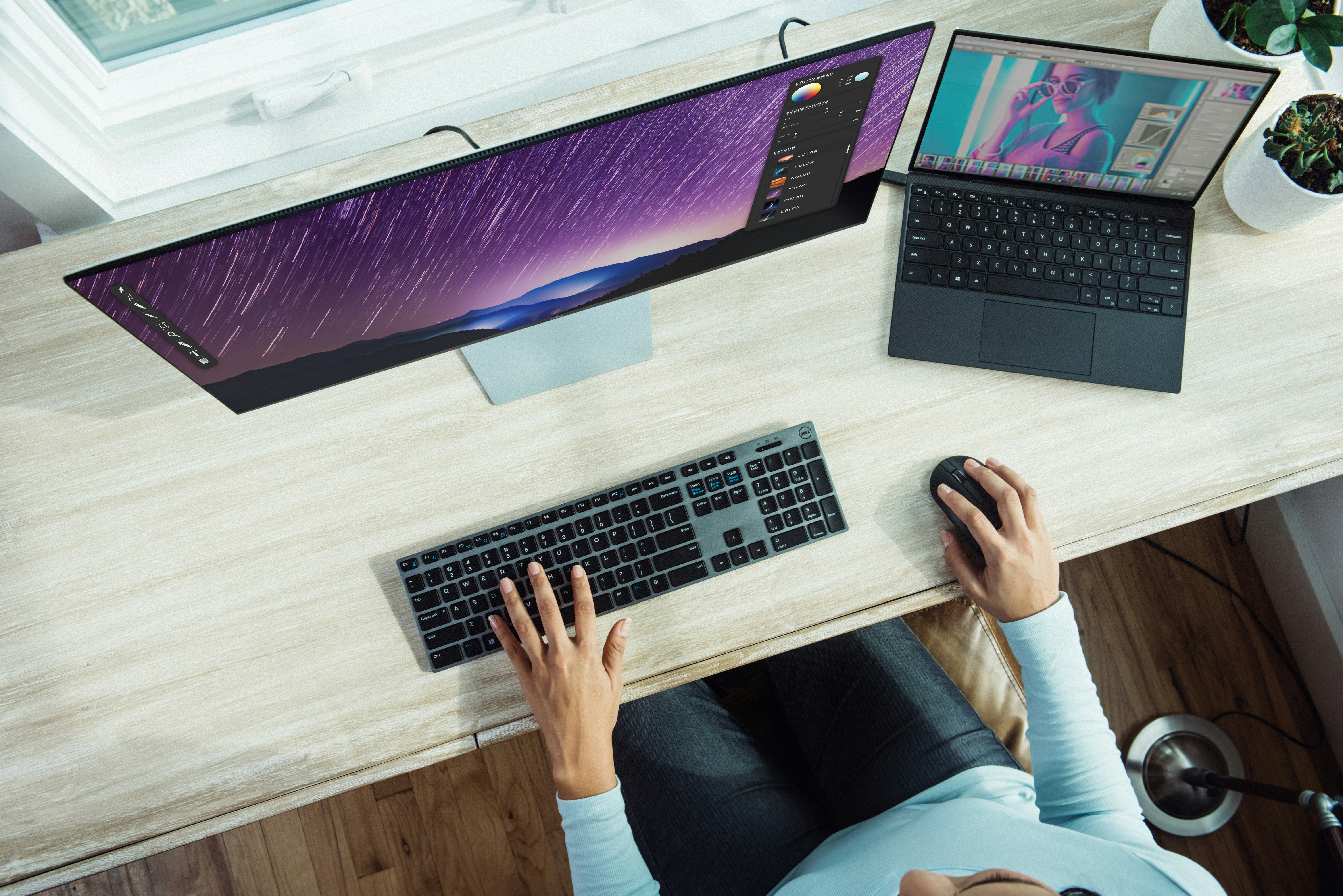 Pessoa trabalhando em um ambiente moderno e bem iluminado. Ela está sentada em frente a uma mesa de madeira clara, com dois monitores e um laptop. O monitor à esquerda exibe gráficos coloridos, enquanto o laptop à direita mostra uma interface de edição de imagem com uma foto de uma mulher. Um vaso com uma planta verde está ao lado do laptop, adicionando um toque natural ao espaço de trabalho.