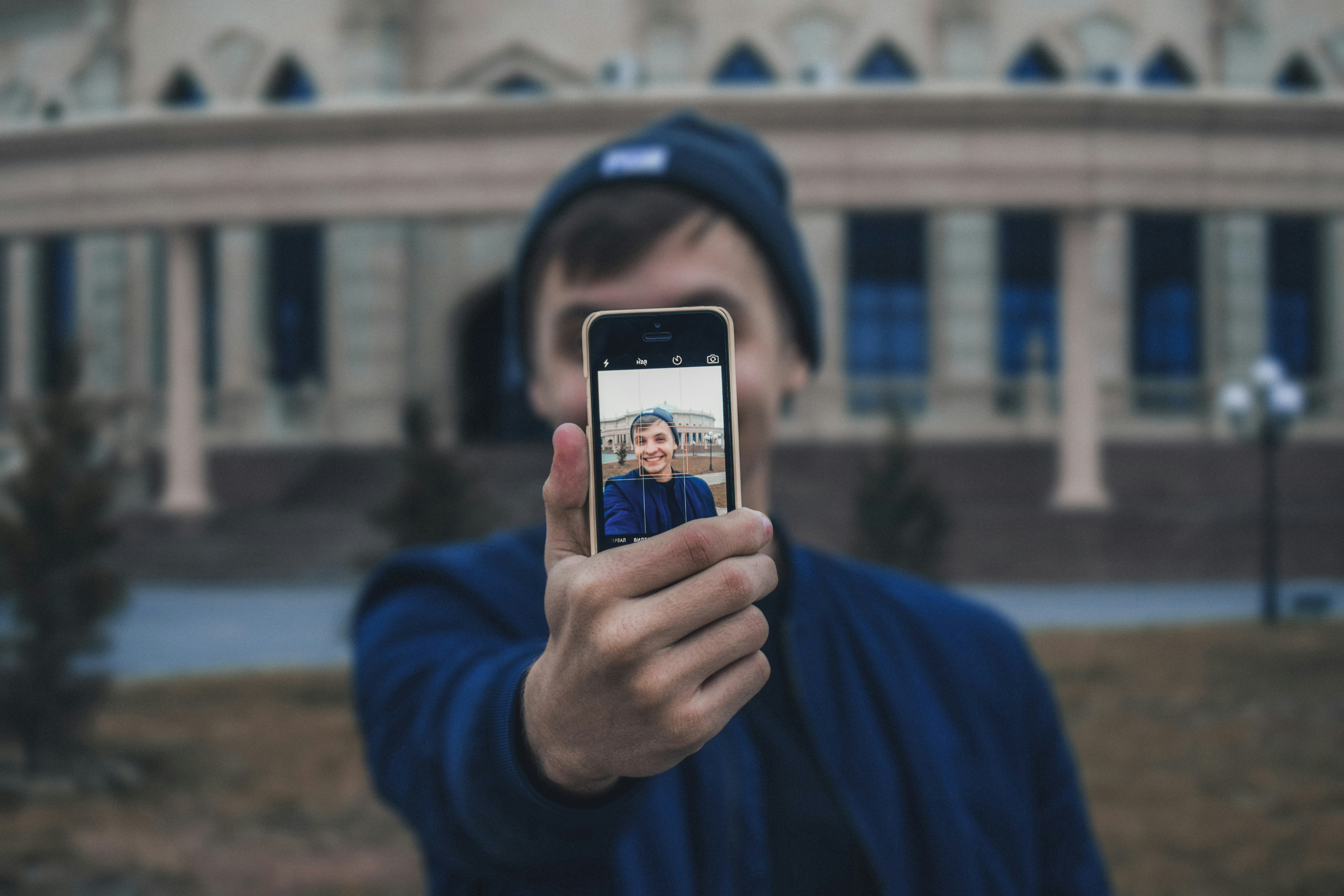 pessoa tirando uma selfie com um prédio elegante ao fundo. O prédio possui múltiplos arcos e janelas, e há árvores sem folhas ao redor. A pessoa está vestindo uma roupa azul e um boné, e o céu parece estar nublado, criando uma atmosfera calma. Representando o controle de ponto digital