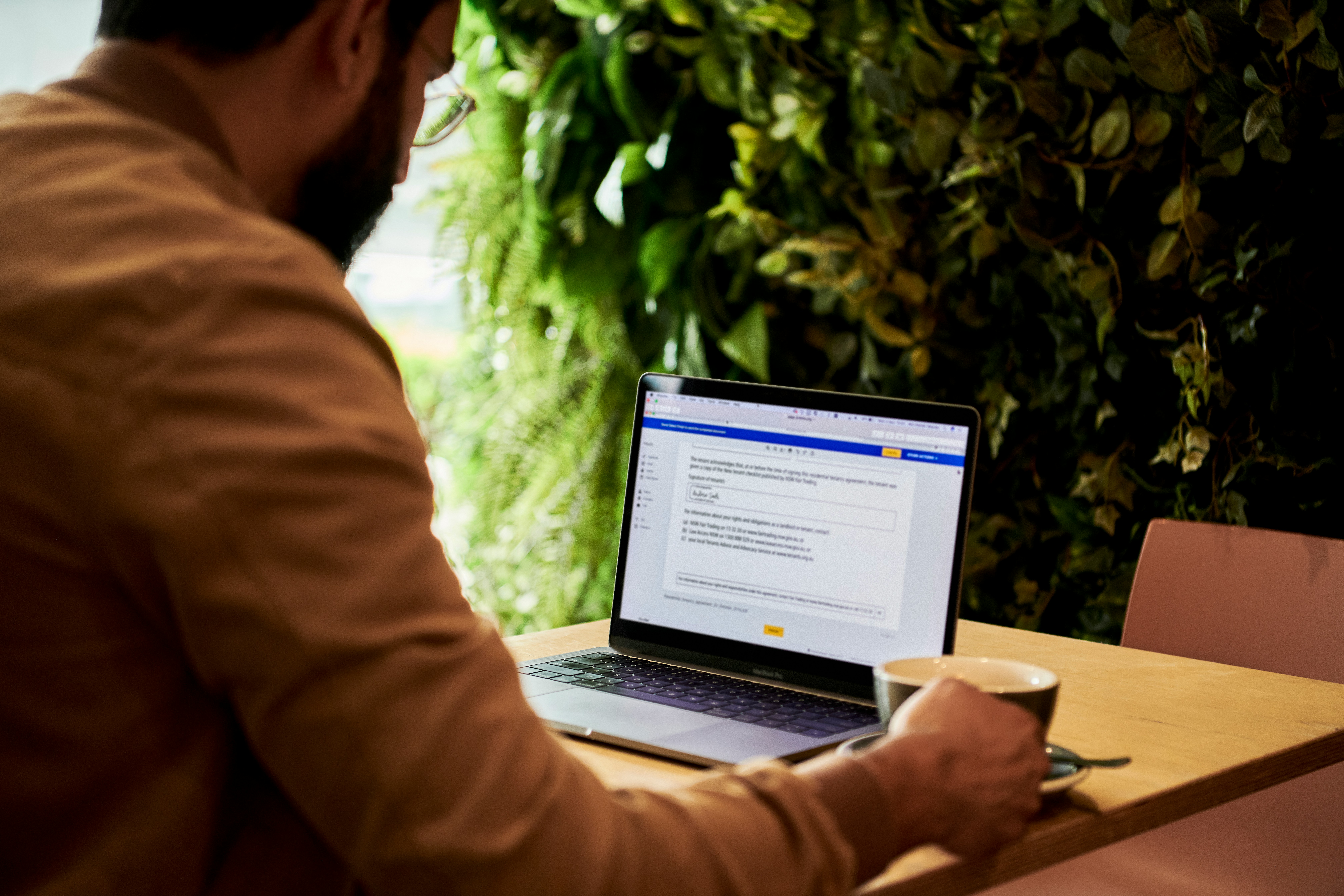Pessoa sentada de costas para a câmera, trabalhando em um laptop sobre uma mesa de madeira. A tela do laptop exibe o que parece ser um documento de texto. Ao lado do laptop, há uma xícara branca que sugere ser de café ou chá. O ambiente tem um tom acolhedor, com iluminação suave e uma parede ao fundo adornada com plantas verdes, o que dá uma sensação de conforto e tranquilidade.