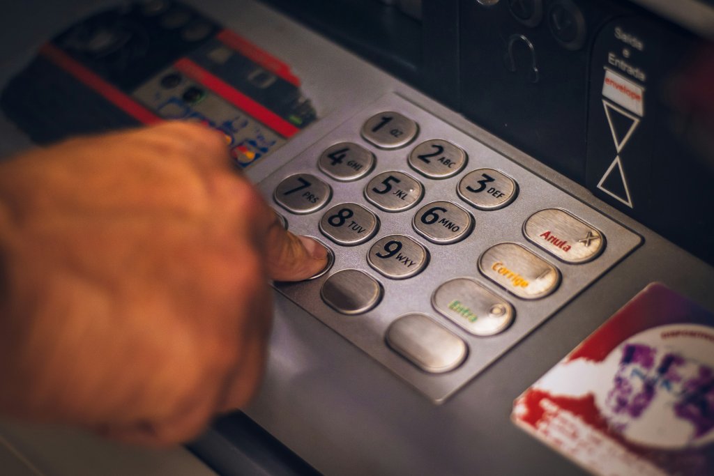 Mão pressionando um botão em um teclado numérico de um caixa eletrônico, com um cartão visível ao lado.
