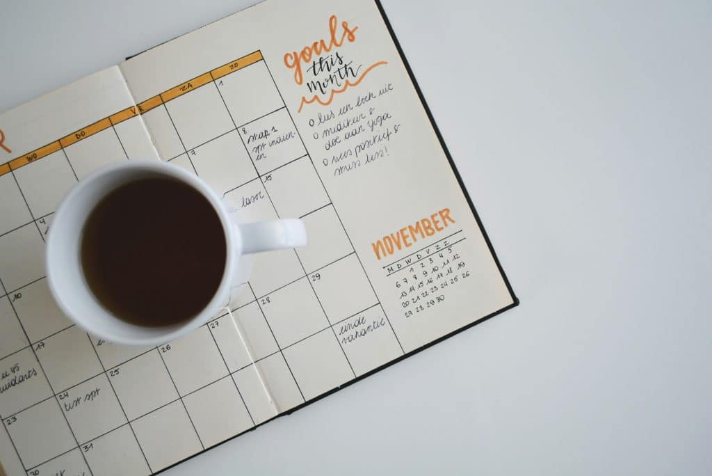 A imagem mostra um planejador aberto na página de novembro, com uma xícara de café colocada em cima dele. O planejador tem seções para metas do mês e datas, e está disposto sobre uma superfície branca.