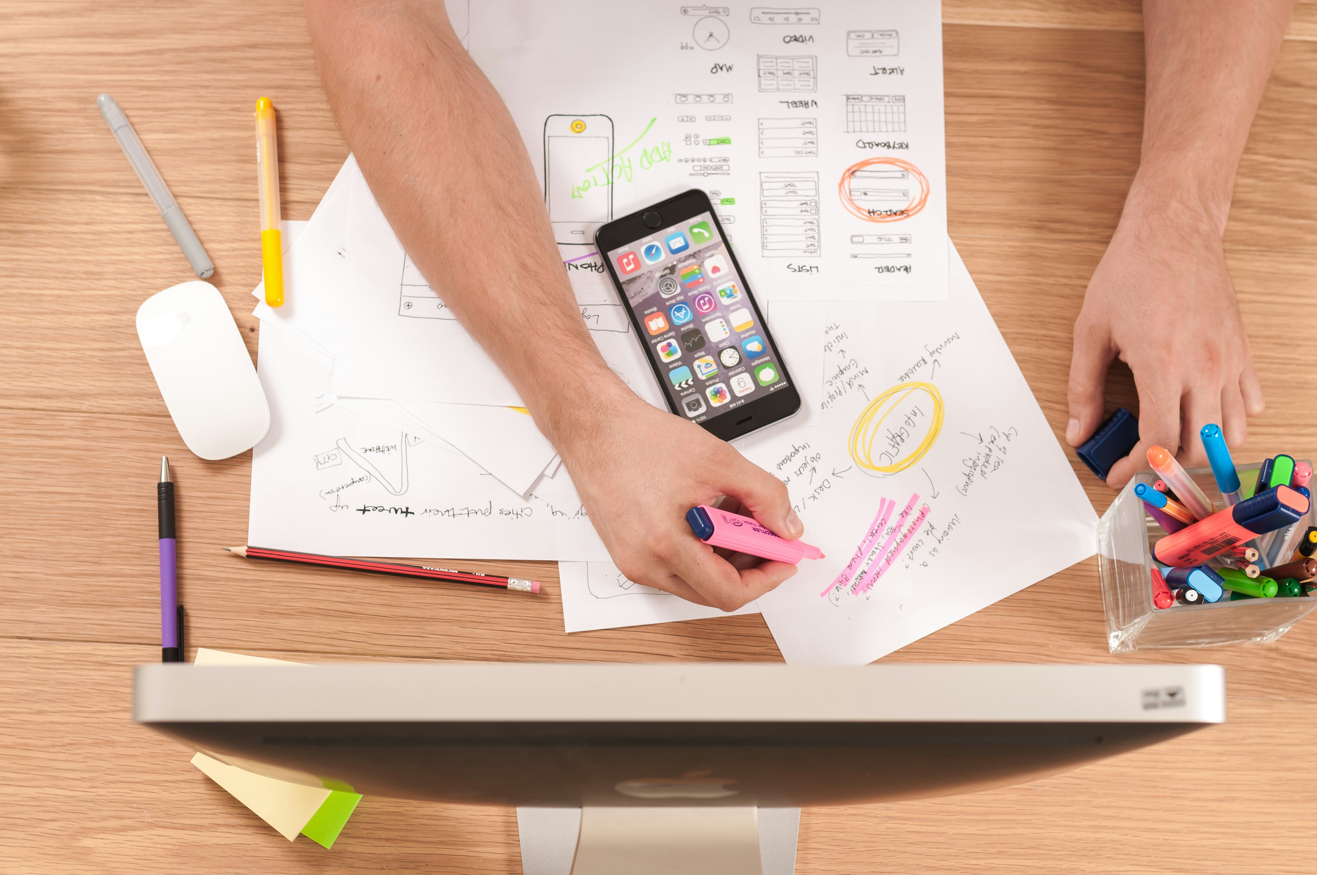 Pessoa destacando informações em um papel com um marcador rosa, enquanto um smartphone e outros itens de escritório estão espalhados sobre a mesa. Os documentos parecem ser esboços ou planos, e a pessoa utiliza vários marcadores coloridos para realçar detalhes.