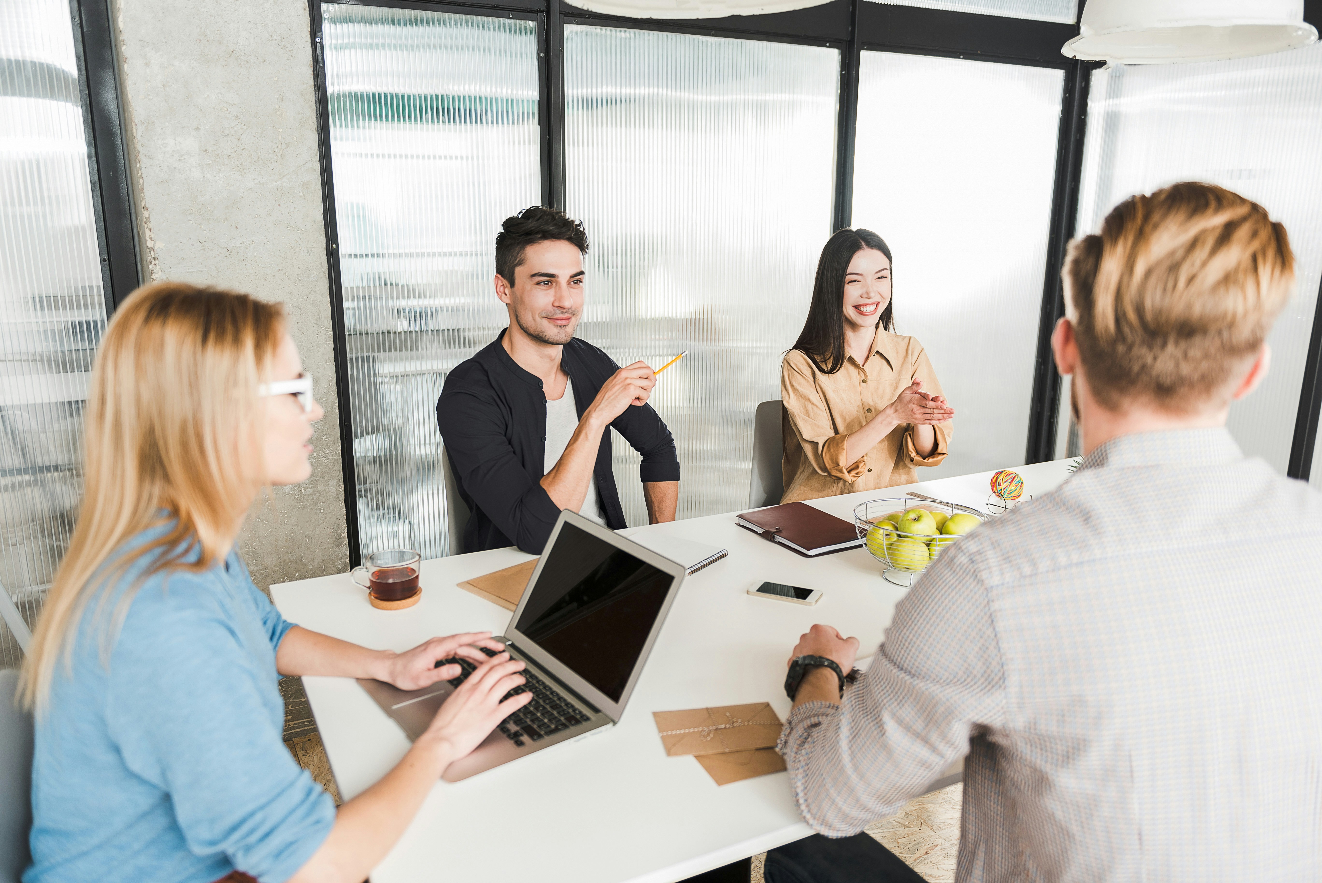quatro pessoas sentadas ao redor de uma mesa em uma sala de reuniões com paredes de vidro. Três das pessoas estão de frente para a câmera. A quarta pessoa está de costas para a câmera. Na mesa, há um laptop, um caderno, algumas canetas e uma tigela de maçãs verdes.