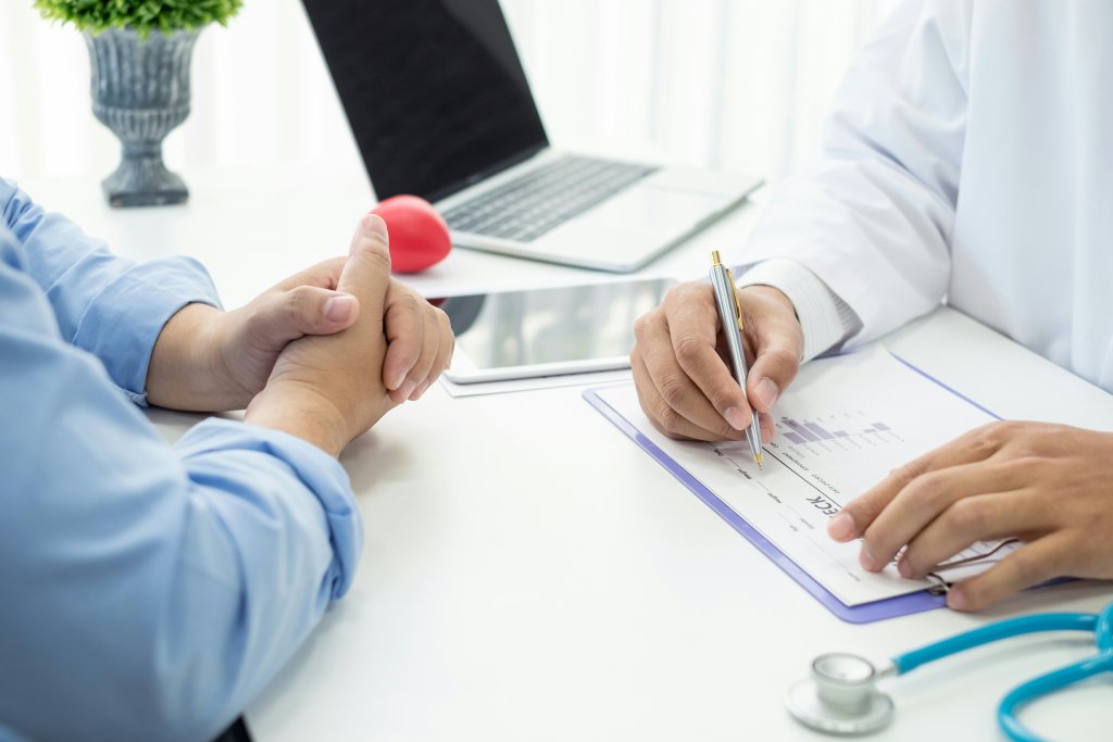 A imagem retrata uma cena de consulta médica. À direita, temos uma pessoa vestindo um jaleco branco, indicando que é um profissional de saúde. Ela segura uma caneta e escreve em um documento que parece estar em uma prancheta. À esquerda, outra pessoa está sentada em frente ao profissional de saúde, com o polegar levantado, possivelmente indicando boa saúde ou concordância. Ao fundo, há um laptop aberto sobre uma mesa e o que parece ser parte de um estetoscópio.