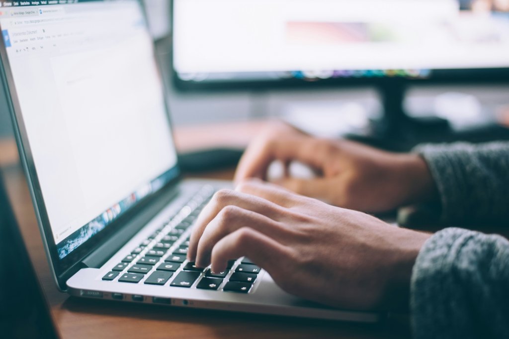 A imagem mostra um close das mãos de uma pessoa digitando em um teclado de laptop. O laptop está sobre uma mesa, e ao fundo, desfocado, parece haver outro monitor ou tela.