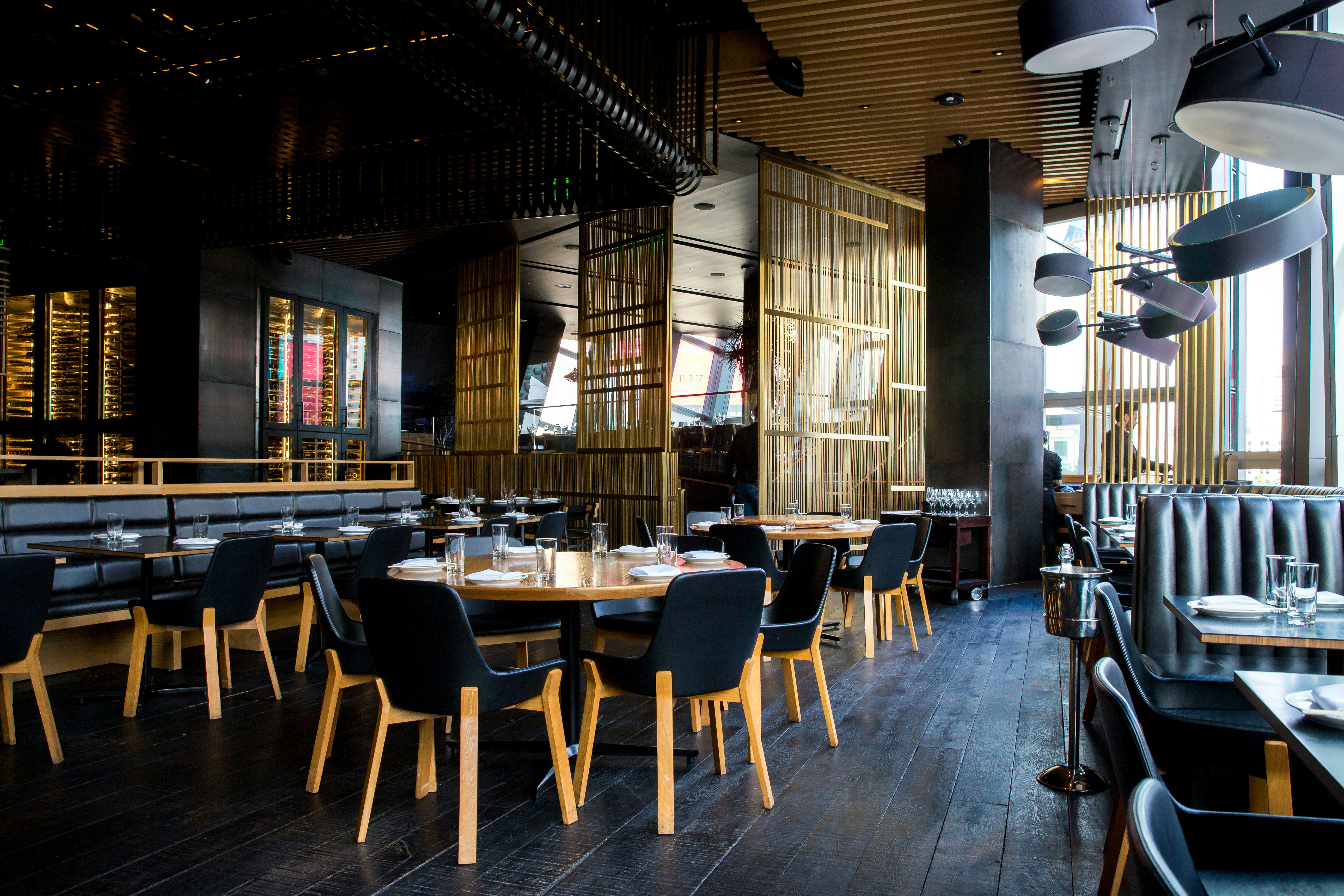 Interior de um restaurante moderno com design sofisticado. O espaço possui teto alto com detalhes em preto e madeira, grandes janelas que permitem a entrada de luz natural e mesas arrumadas para o serviço. Destaque para as divisórias verticais douradas que criam espaços semi-privativos dentro do ambiente amplo e aberto. A paleta de cores é predominantemente preta, dourada e tons de madeira, conferindo uma atmosfera acolhedora e elegante.