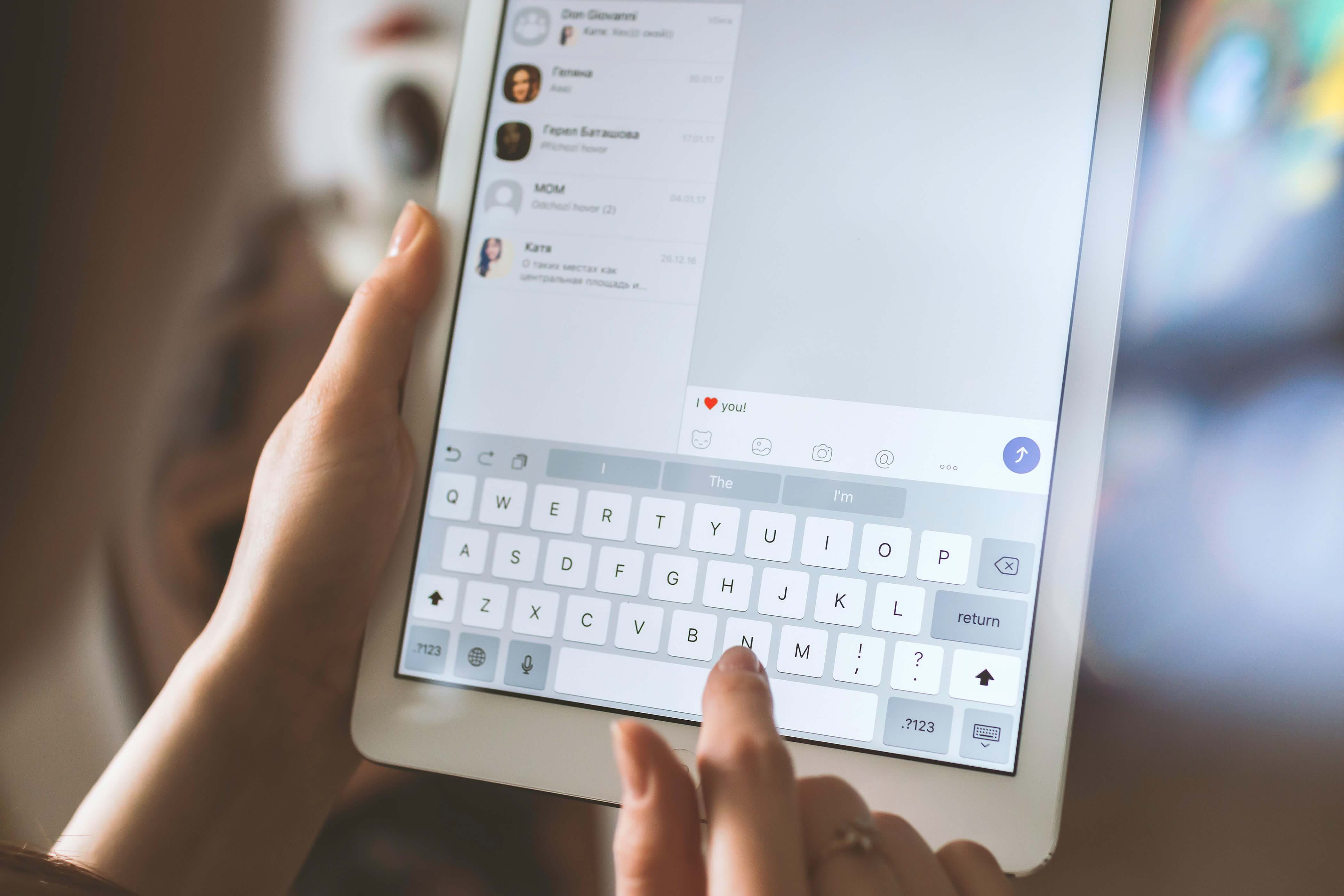 Pessoa segurando um tablet ou iPad, digitando uma mensagem. A tela do dispositivo exibe um aplicativo de mensagens, com uma lista de conversas à esquerda e a caixa de texto ativa à direita. O teclado virtual está visível na parte inferior da tela, com botões de emojis e opções adicionais ao lado da barra de espaço. O fundo está levemente desfocado, dando foco total ao dispositivo e à interação de digitação.