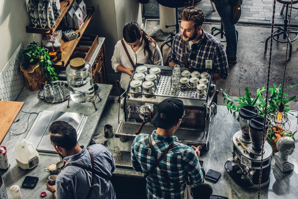 Visão aérea de uma cena em uma cafeteria. Quatro pessoas estão ao redor de um balcão de café equipado com máquinas de espresso e vários acessórios para fazer café. Uma pessoa parece estar preparando uma bebida, outra está interagindo com o que parece ser um cliente, e as outras duas estão envolvidas em atividades separadas. O ambiente tem uma decoração de estilo industrial com paredes de tijolos expostos e luminárias pendentes.