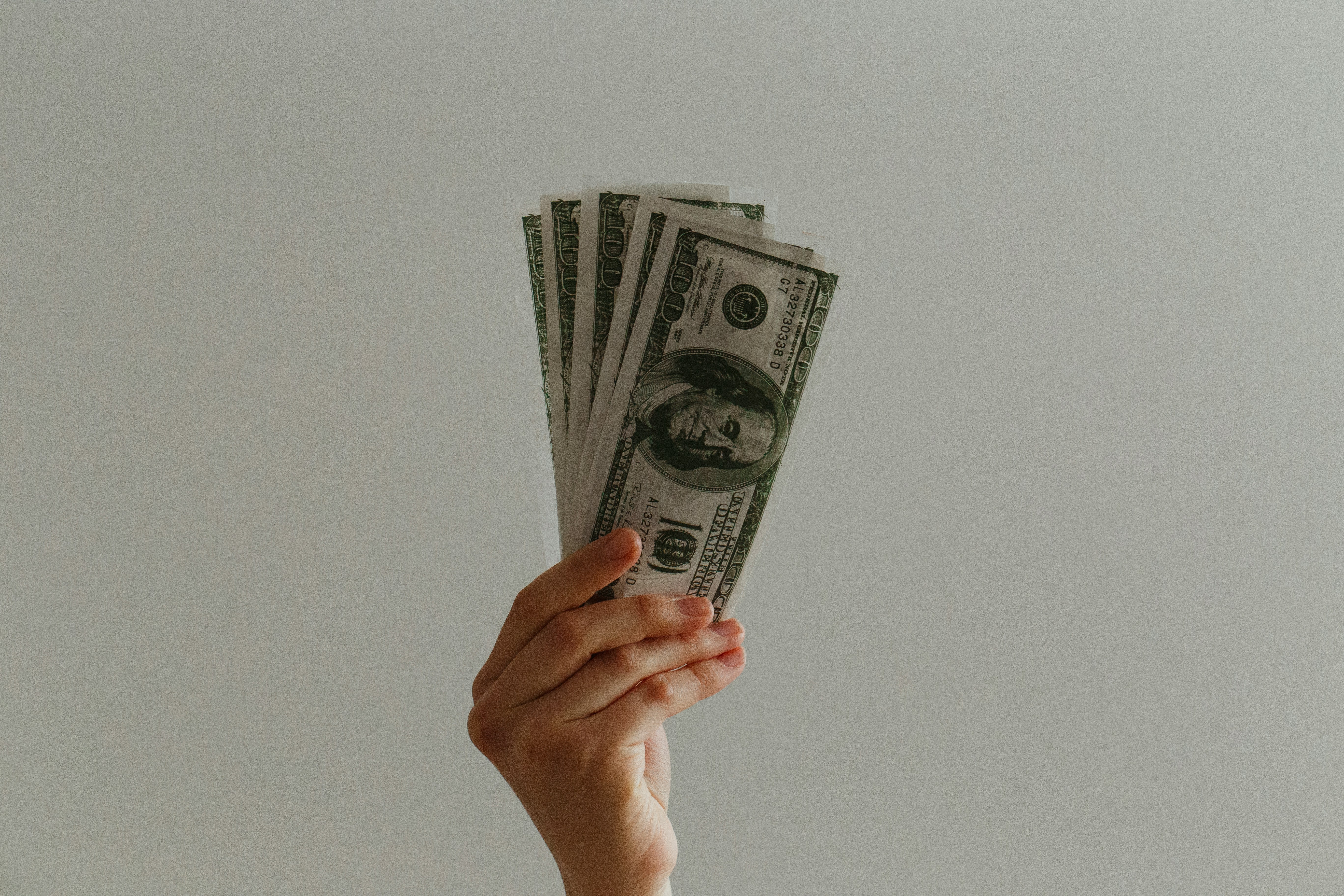 mão segurando um leque de notas de dólar dos Estados Unidos em um fundo claro. As denominações das notas não estão totalmente visíveis, mas é possível identificar que são notas de 1 dólar.