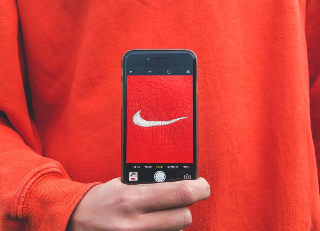 Pessoa segurando um iPhone, que exibe a tela da câmera com uma imagem do logo da Nike em um fundo vermelho, correspondendo à cor da roupa que a pessoa está vestindo.  O registro visual captura a sinergia entre tecnologia e marcas populares.