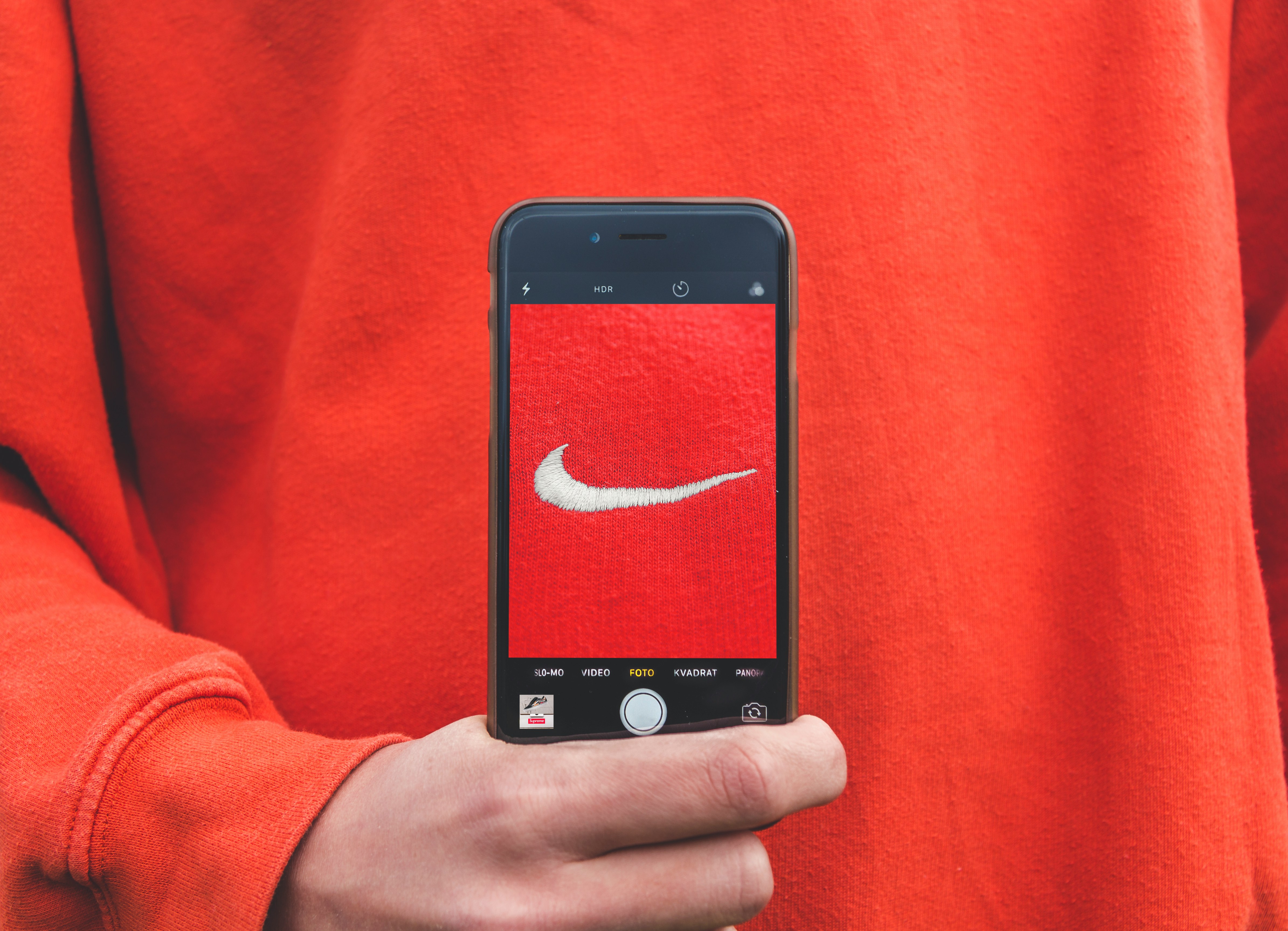 Pessoa segurando um iPhone, que exibe a tela da câmera com uma imagem do logo da Nike em um fundo vermelho, correspondendo à cor da roupa que a pessoa está vestindo. O registro visual captura a sinergia entre tecnologia e marcas populares.