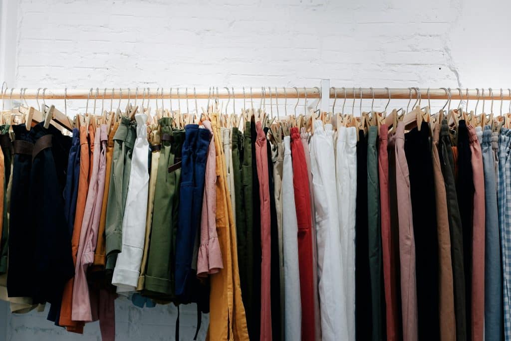 Várias calças penduradas ordenadamente em cabides. Elas vêm em uma variedade de cores, incluindo preto, verde, amarelo, vermelho e azul. Os cabides são de madeira e estão suspensos em um trilho fixado na parede branca ao fundo.