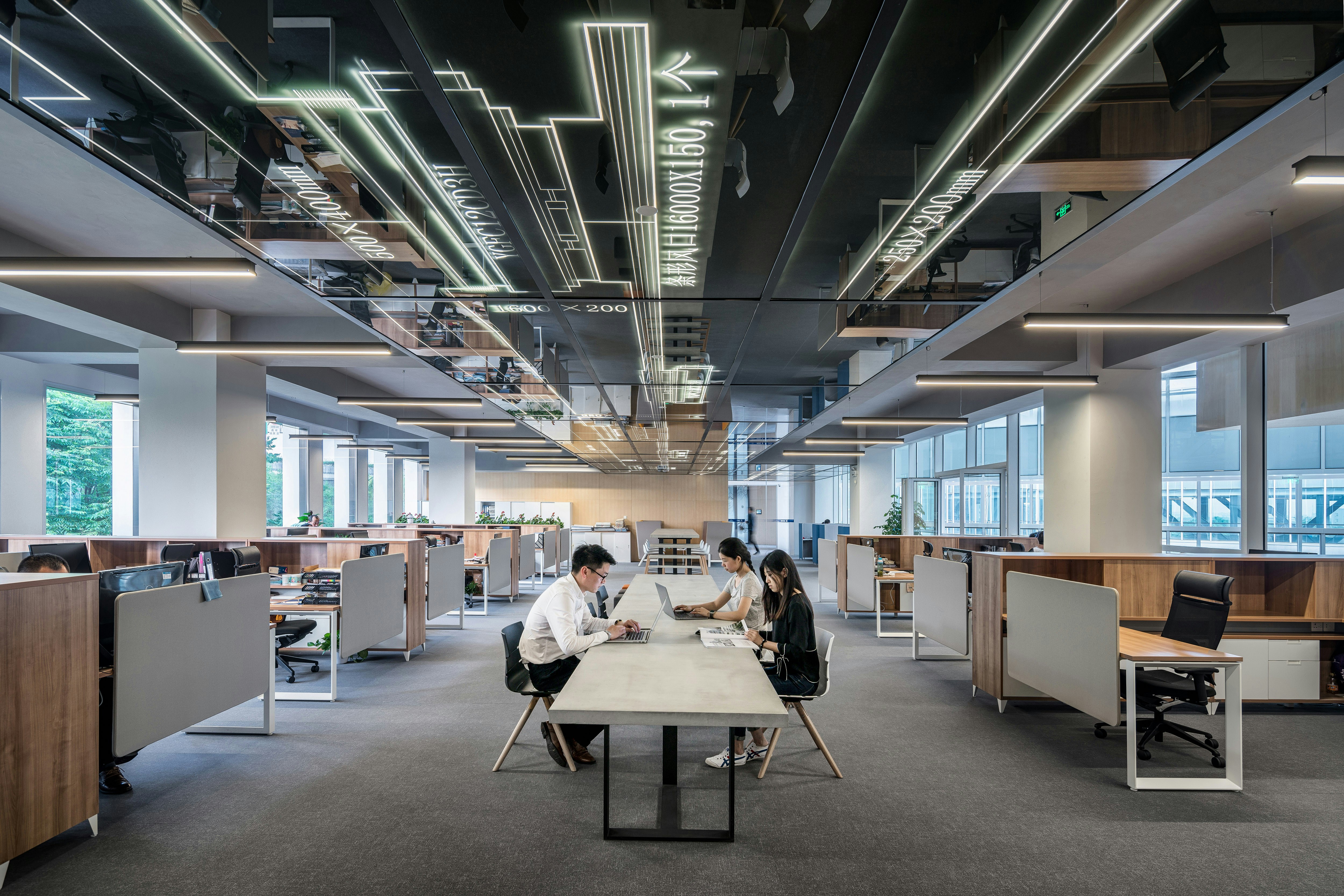 escritório moderno com um grupo de três pessoas sentadas em uma mesa central, aparentemente envolvidas em discussão ou trabalho. O escritório é espaçoso e bem iluminado, com luz natural vindo de grandes janelas à direita. O design de interiores apresenta elementos contemporâneos, como móveis elegantes, estações de trabalho individuais com computadores desktop e um teto chamativo com luminárias geométricas. A presença de vegetação pode ser vista do lado de fora das janelas, adicionando um toque de natureza ao ambiente.