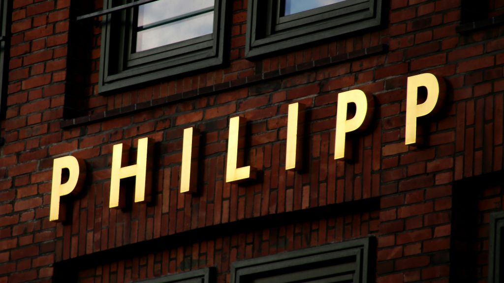 nome “PHILIPP” em letras grandes e iluminadas, fixadas na parede exterior de um edifício de tijolos vermelhos com janelas escuras.  A combinação entre as letras brilhantes e os elementos arquitetônicos escuros cria um contraste interessante