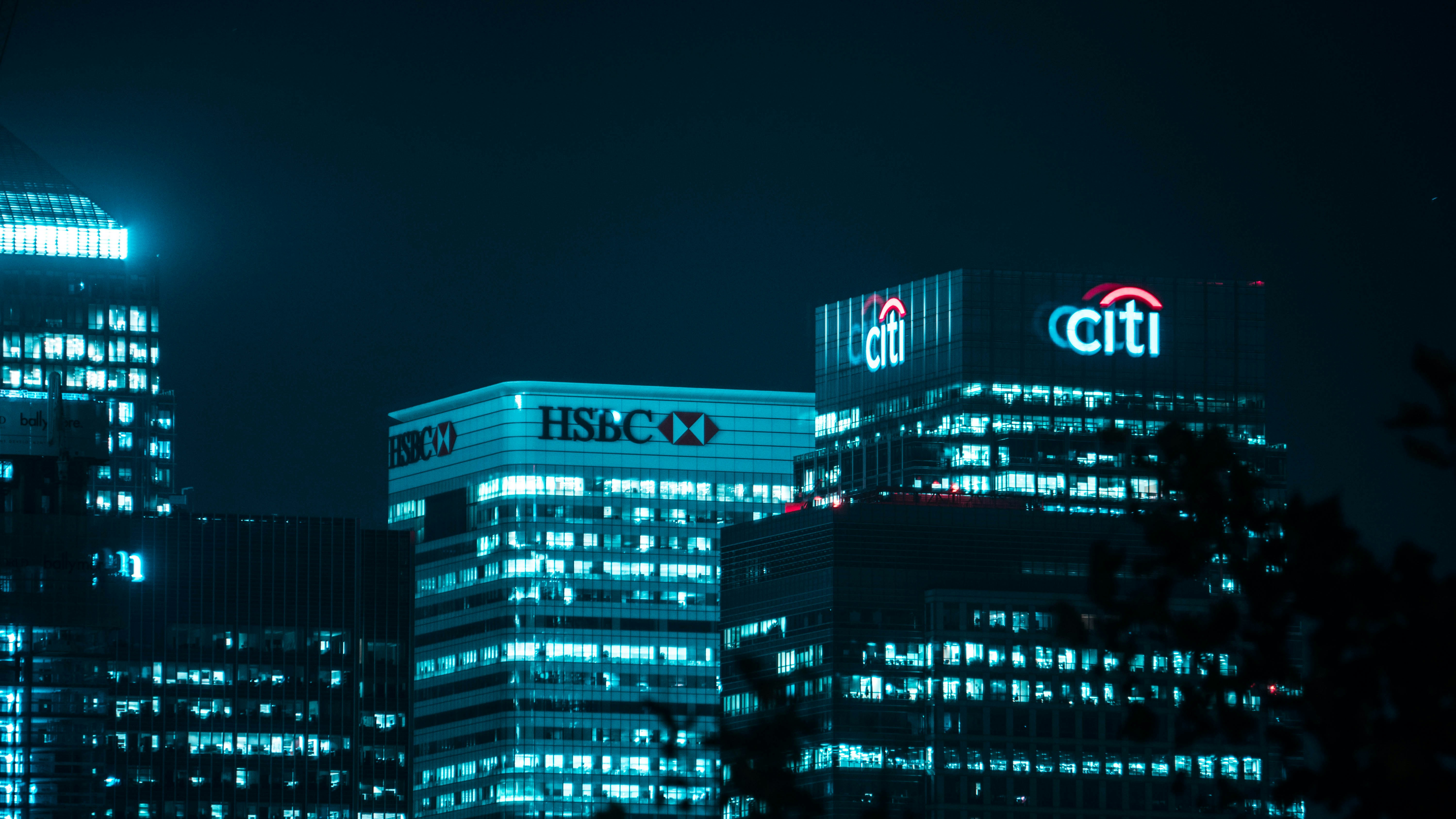 Edifícios iluminados à noite, com destaque para os logotipos luminosos do HSBC e do Citi.