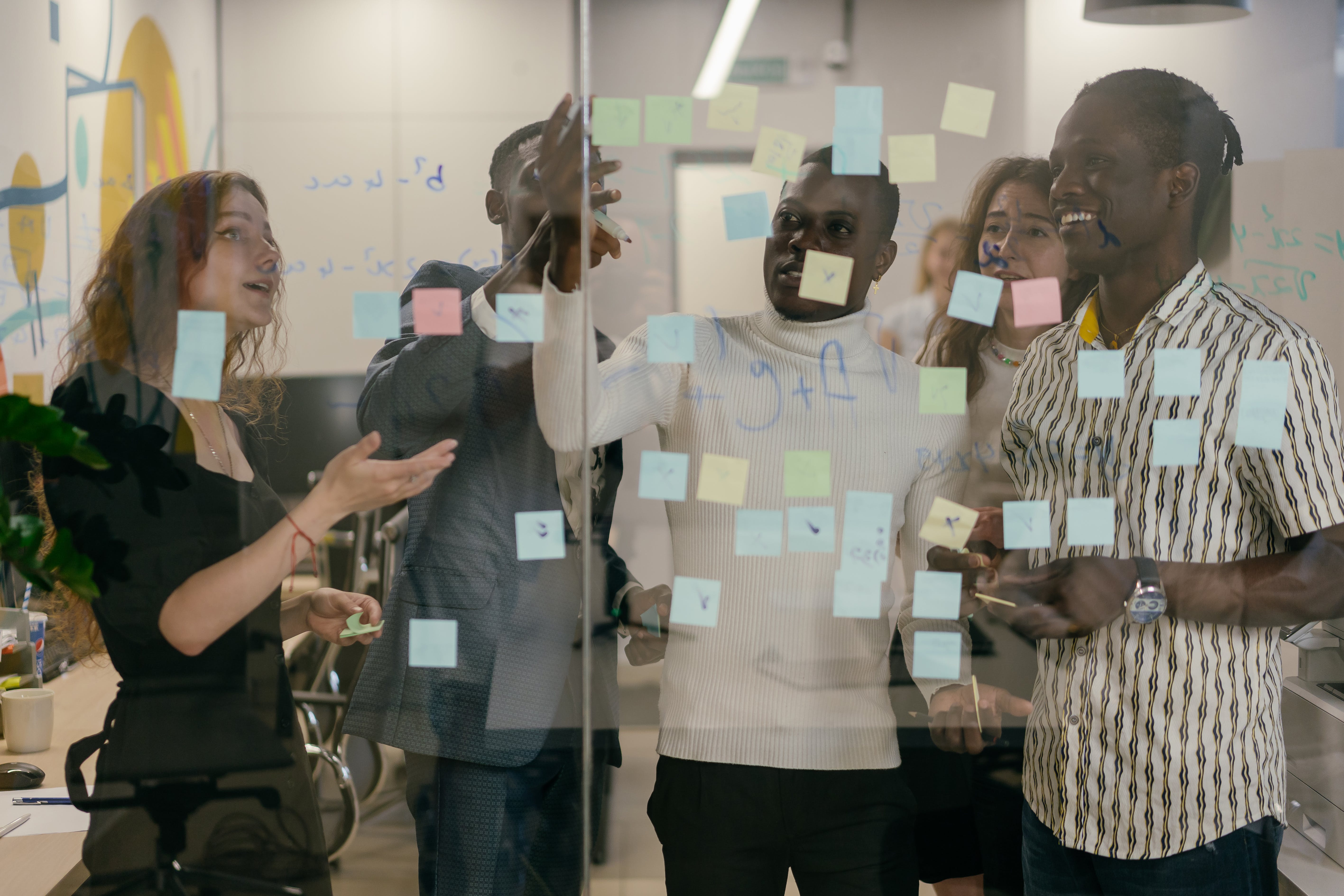 A imagem mostra várias pessoas em uma sala, interagindo perto de uma parede de vidro coberta com post-its coloridos. As faces das pessoas estão obscurecidas para manter a privacidade.
Várias pessoas estão reunidas em um ambiente interno, possivelmente um escritório ou sala de reuniões. A parede de vidro ao fundo está coberta com post-its coloridos, indicando uma sessão de brainstorming ou planejamento. Algumas palavras e desenhos são visíveis nos post-its, mas não são claramente legíveis. As pessoas parecem estar em uma discussão animada ou colaborativa, embora suas emoções específicas não possam ser determinadas. O ambiente é moderno e bem iluminado, com outras salas visíveis ao fundo através da parede de vidro.