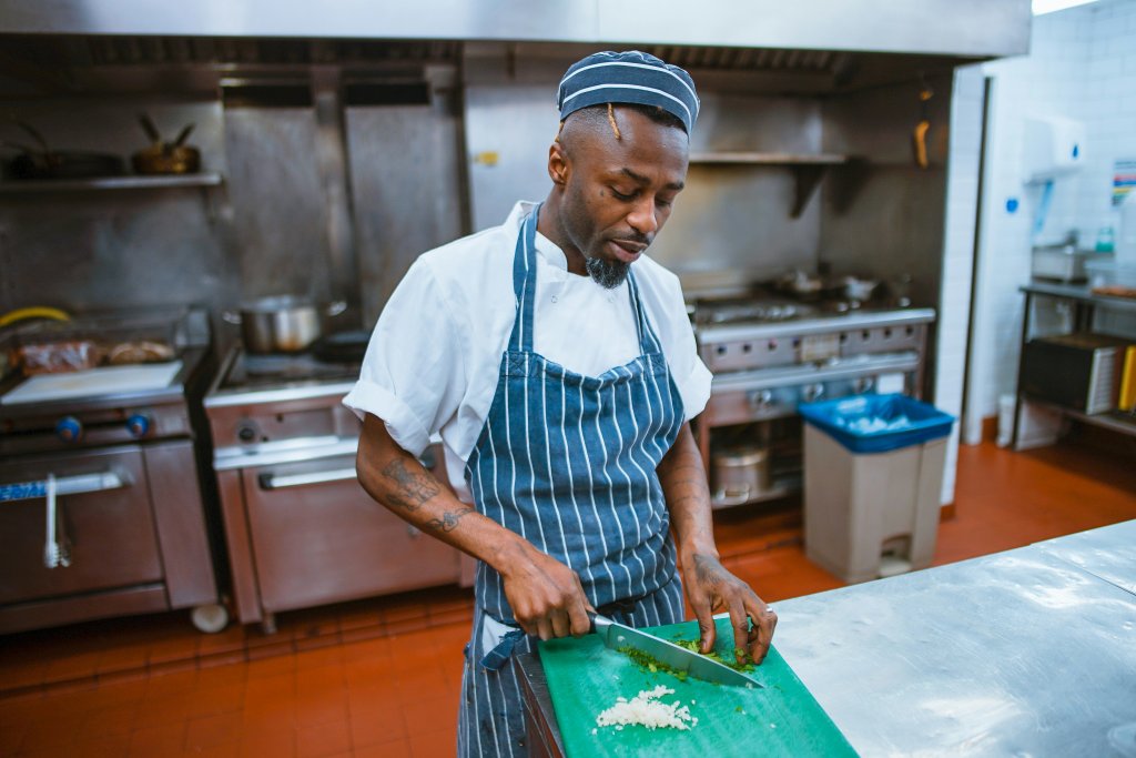 Pessoa vestindo um uniforme de chef, composto por um casaco branco e um avental listrado azul, em uma cozinha comercial. A pessoa está usando uma faca para cortar um ingrediente em uma tábua de corte verde, colocada sobre uma bancada de aço inoxidável. Ao fundo, há vários equipamentos de cozinha, incluindo fogões e fornos.